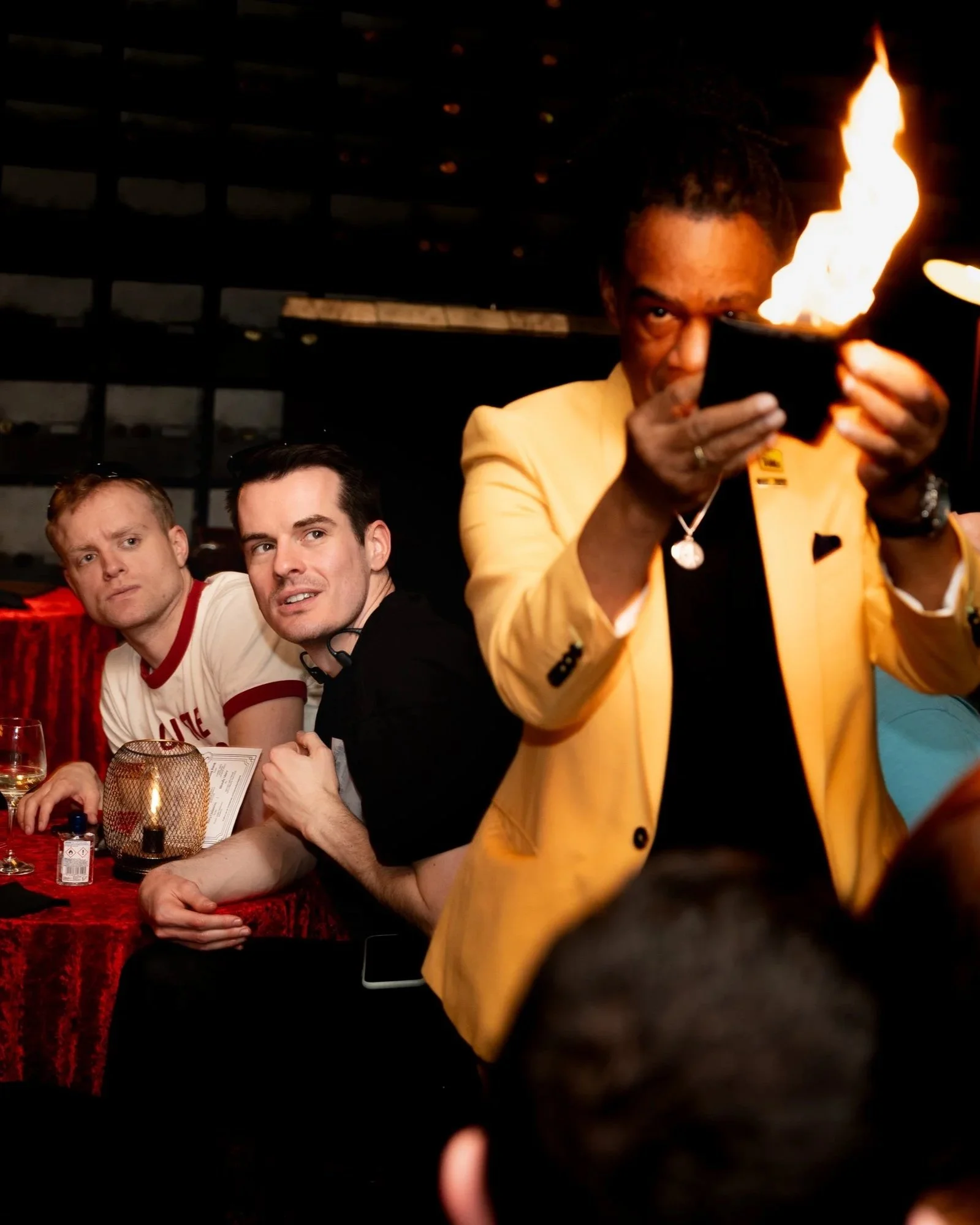 A magician in a yellow jacket holding a flaming wallet while two guests look on