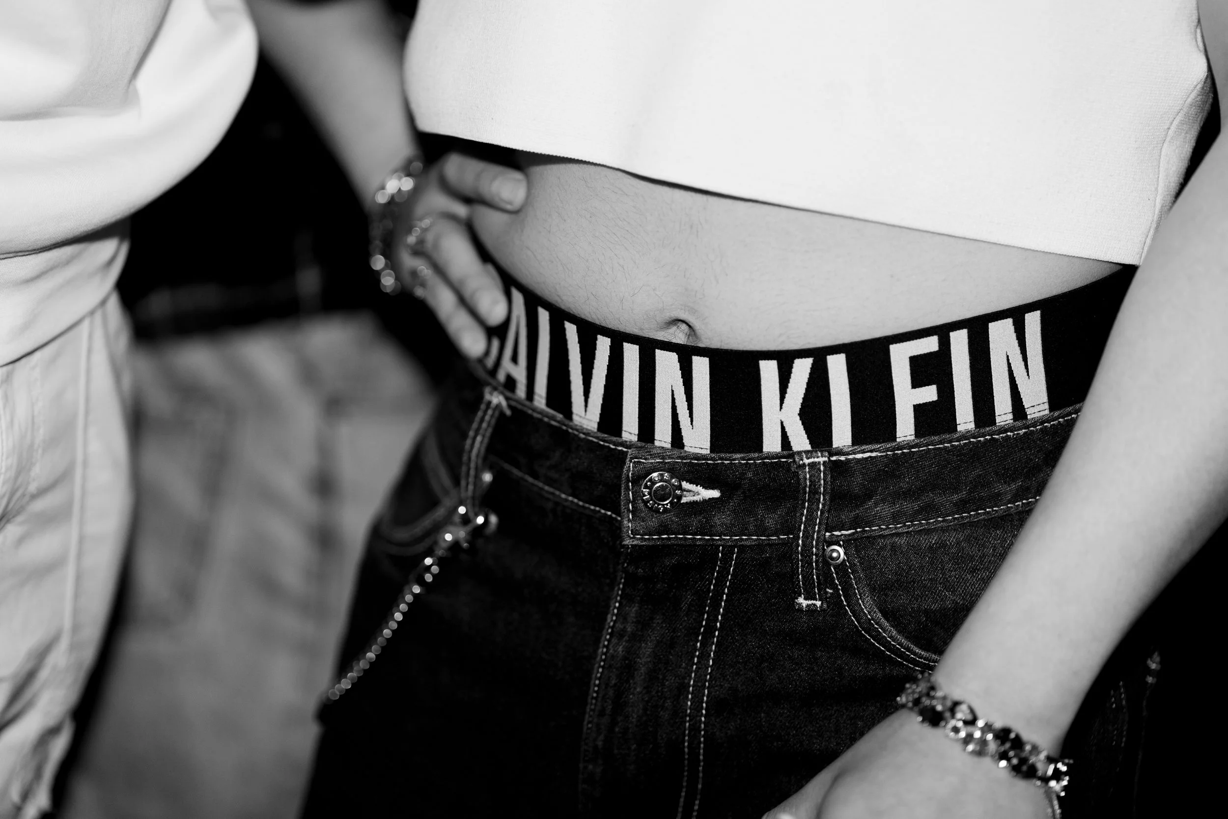 Close-up of a woman's midsection showing her wearing a white crop top, black Calvin Klein underwear, and dark jeans with a chain bracelet on her wrist.