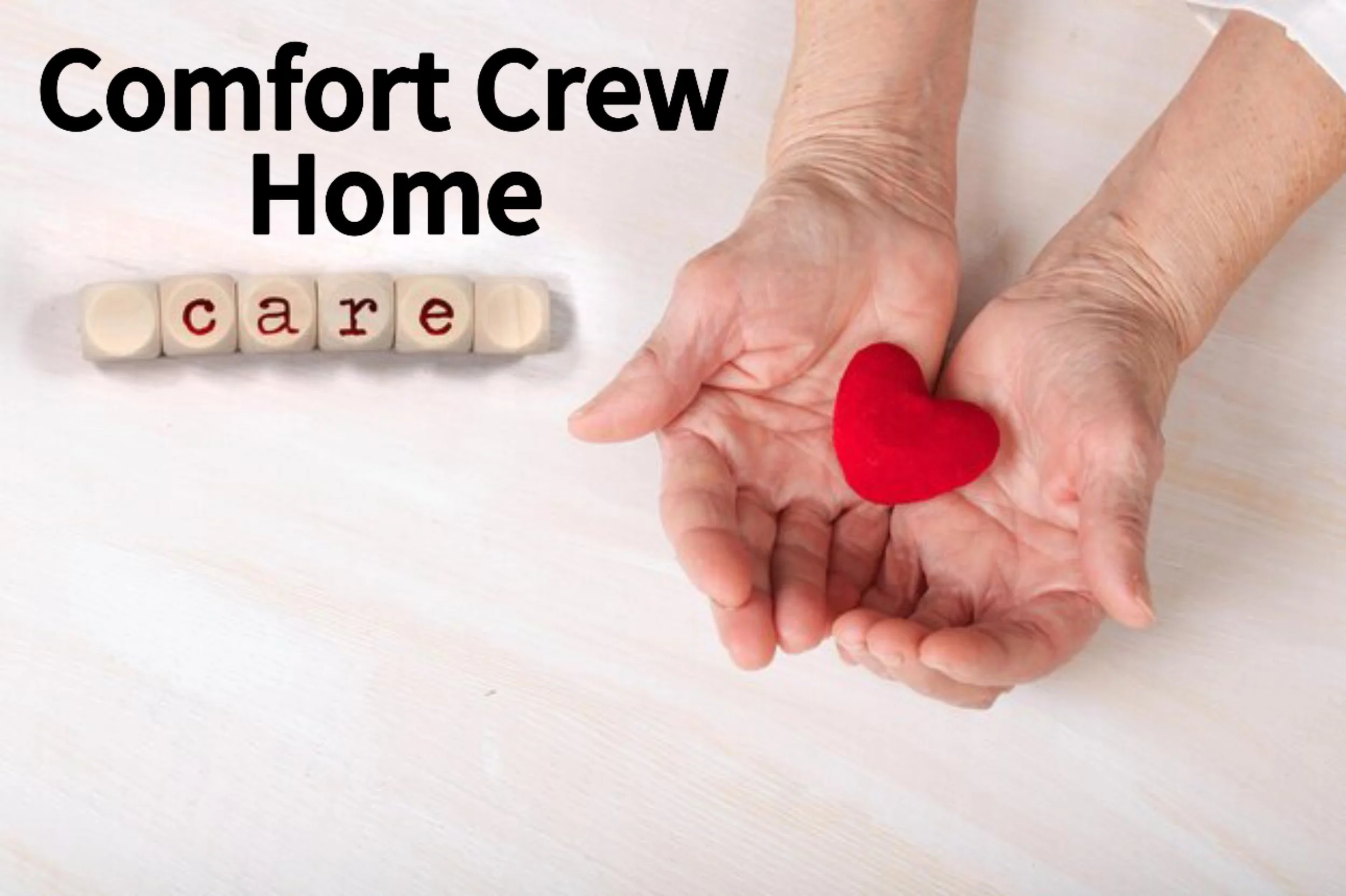Elderly hands holding a small red heart-shaped object on a white surface, with the words 'Comfort Crew Home' and the word 'care' spelled out with wooden letter blocks.