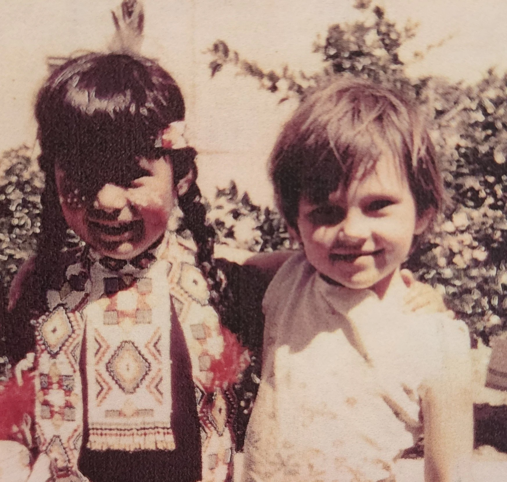 Two children, a girl with braided hair and a boy, smiling outdoors with bushes in the background.