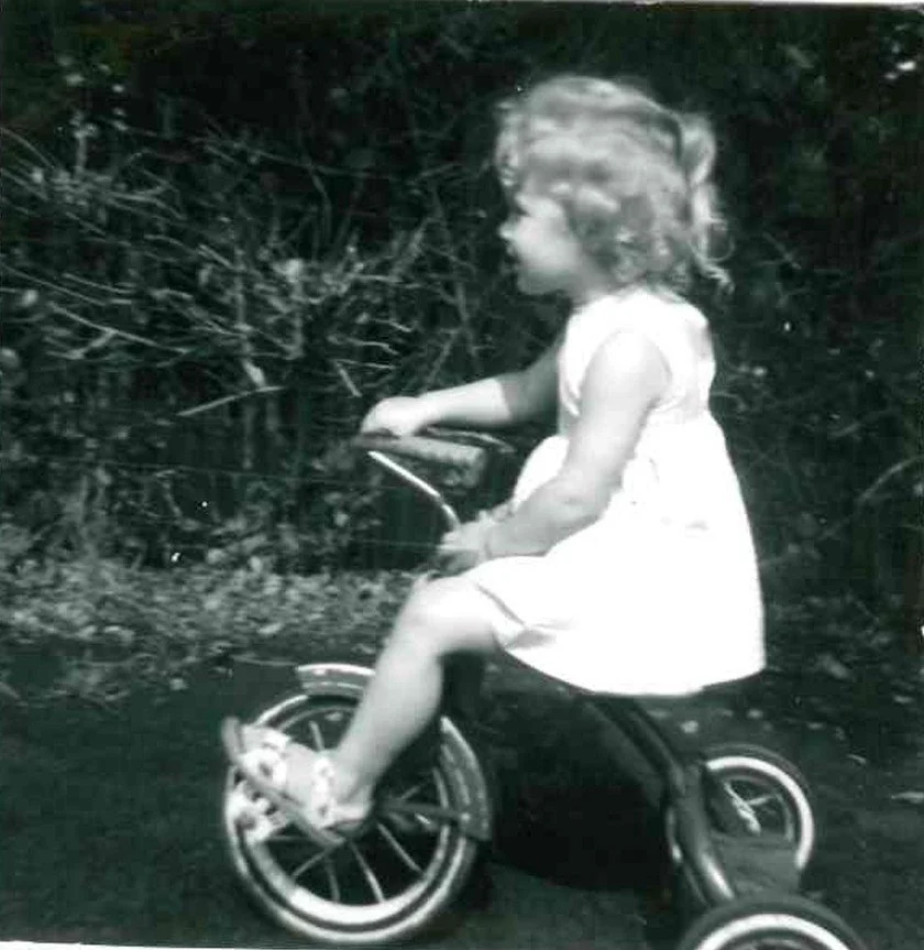 A young girl in a white dress riding a small tricycle outdoors.