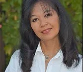 A woman with long black hair, wearing a white shirt, standing outdoors with green trees in the background.