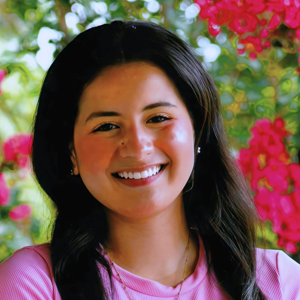A young woman smiling outdoors with pink and green flowering plants in the background.