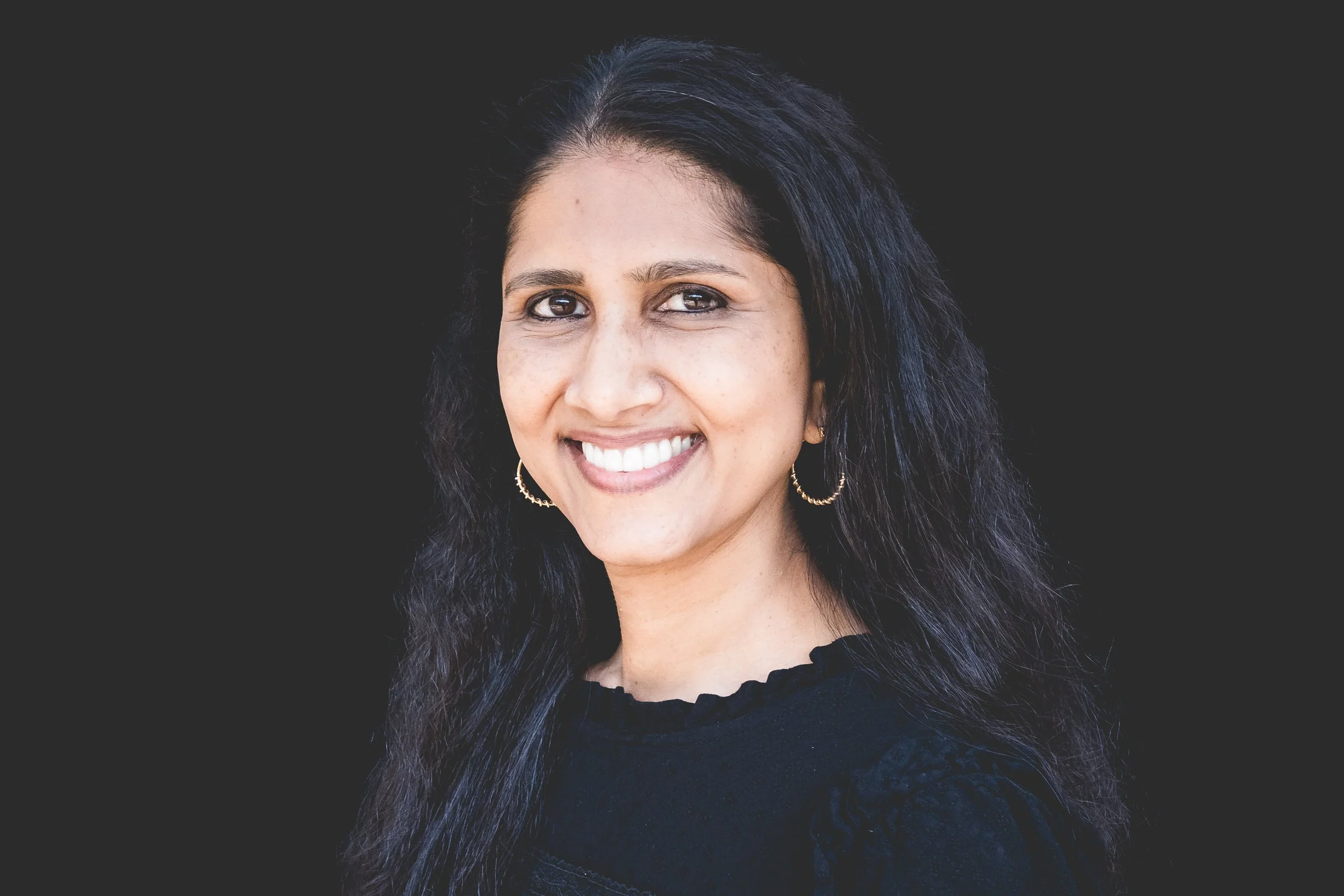 Portrait of a smiling woman with long dark hair, wearing hoop earrings and a black top, against a dark background.