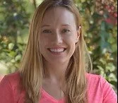 A smiling woman with long blonde hair wearing a pink top, standing outdoors with green foliage in the background.