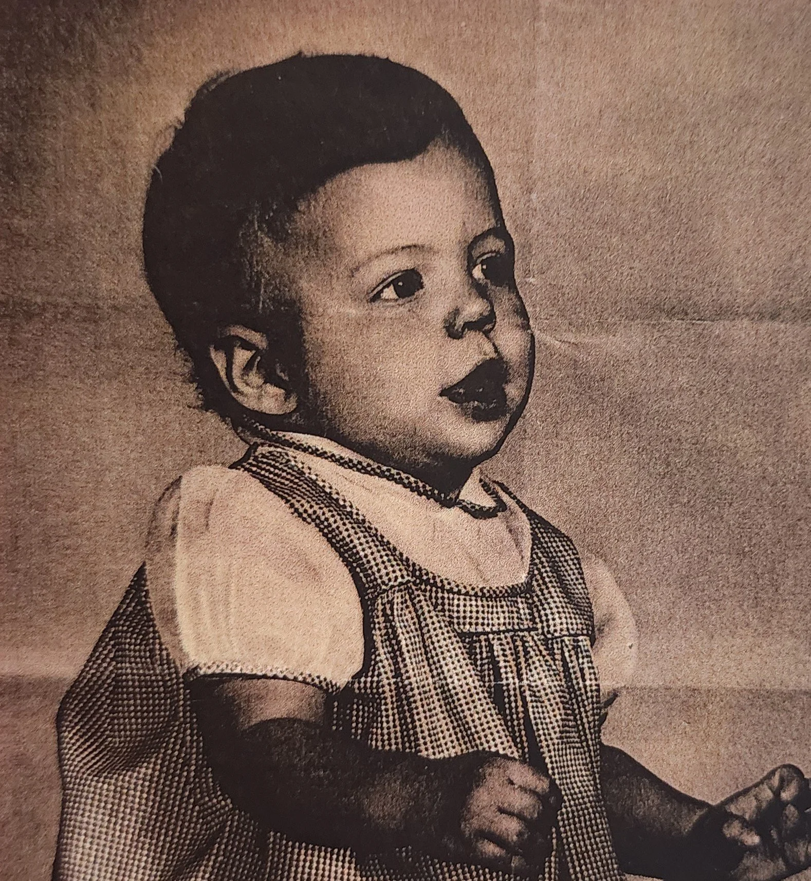 A vintage black and white photograph of a young child with short hair, wearing a checkered dress with puffed sleeves, and holding a small object in her hand.