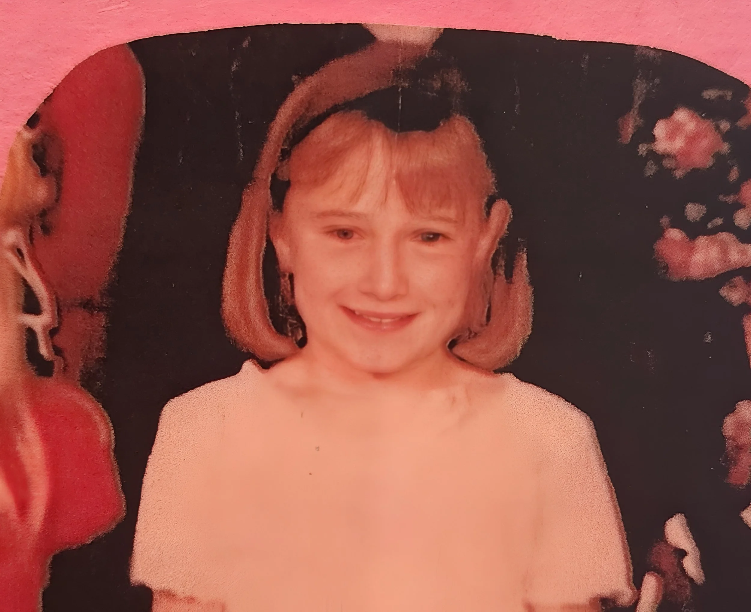 A young boy with blonde hair smiling, dressed in a light pink shirt, with a dark background and some pink flowers.