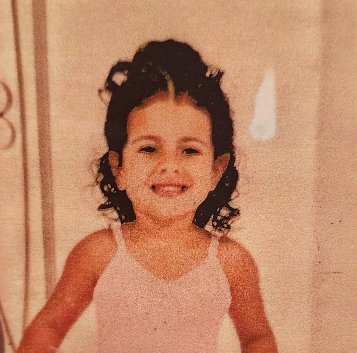 A young girl smiling at the camera, wearing a light-colored sleeveless top, with curly hair styled in pigtails, standing in a room with a beige wall and a white wall sconce in the background.