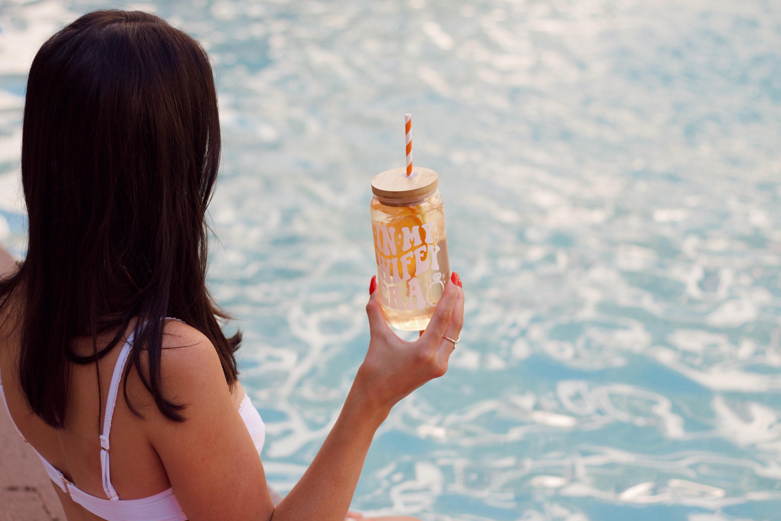 A woman with brunette hair sitting by the water at the beach, holding a glass jar with a straw, decorated with text and imagery, drinking a beverage.