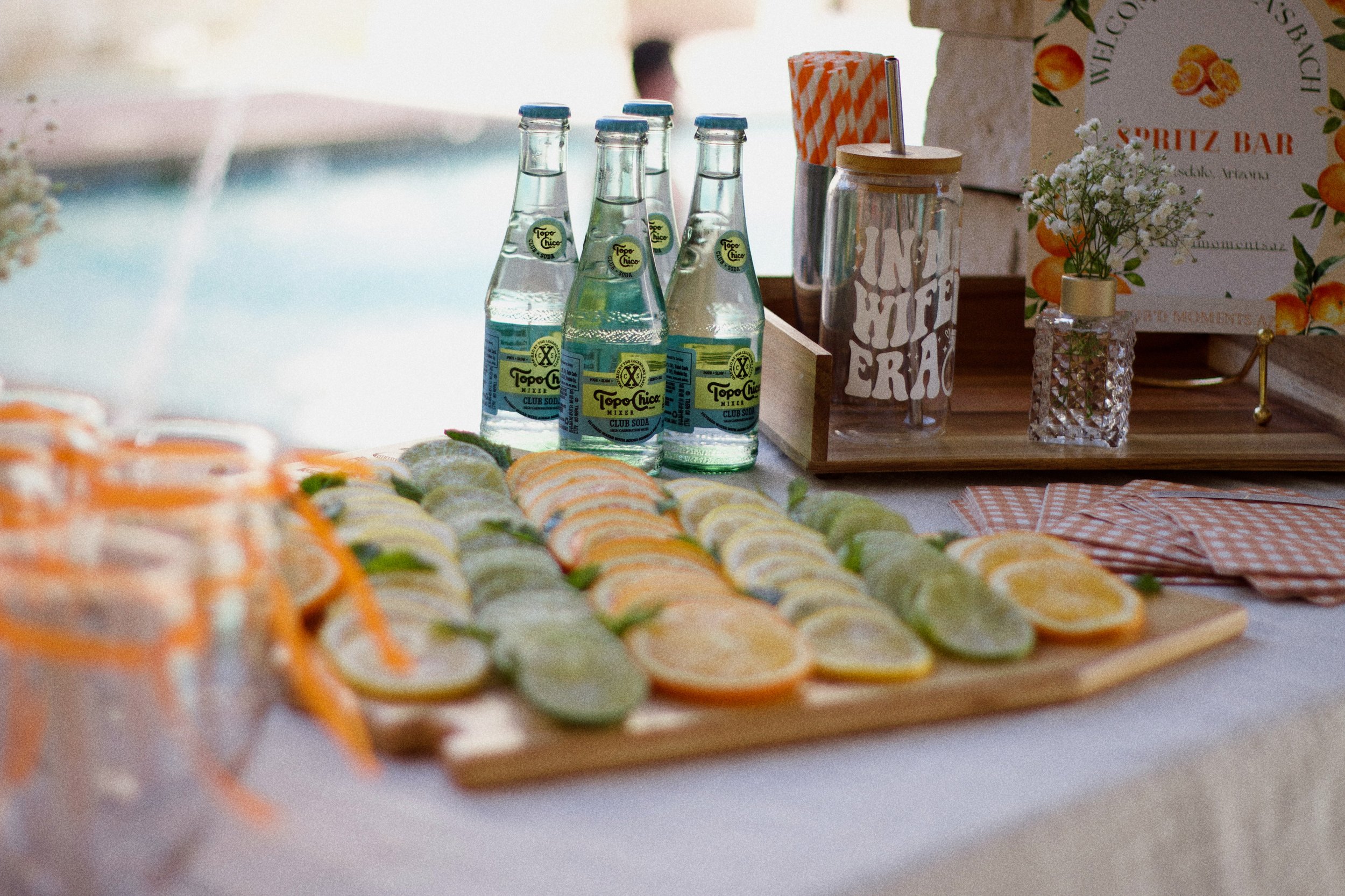 A table decorated with citrus slices, bottles of Topo Chico soda, striped straws, a jar with a straw, a small vase with white flowers, and a sign promoting a spritz bar at a party or event.