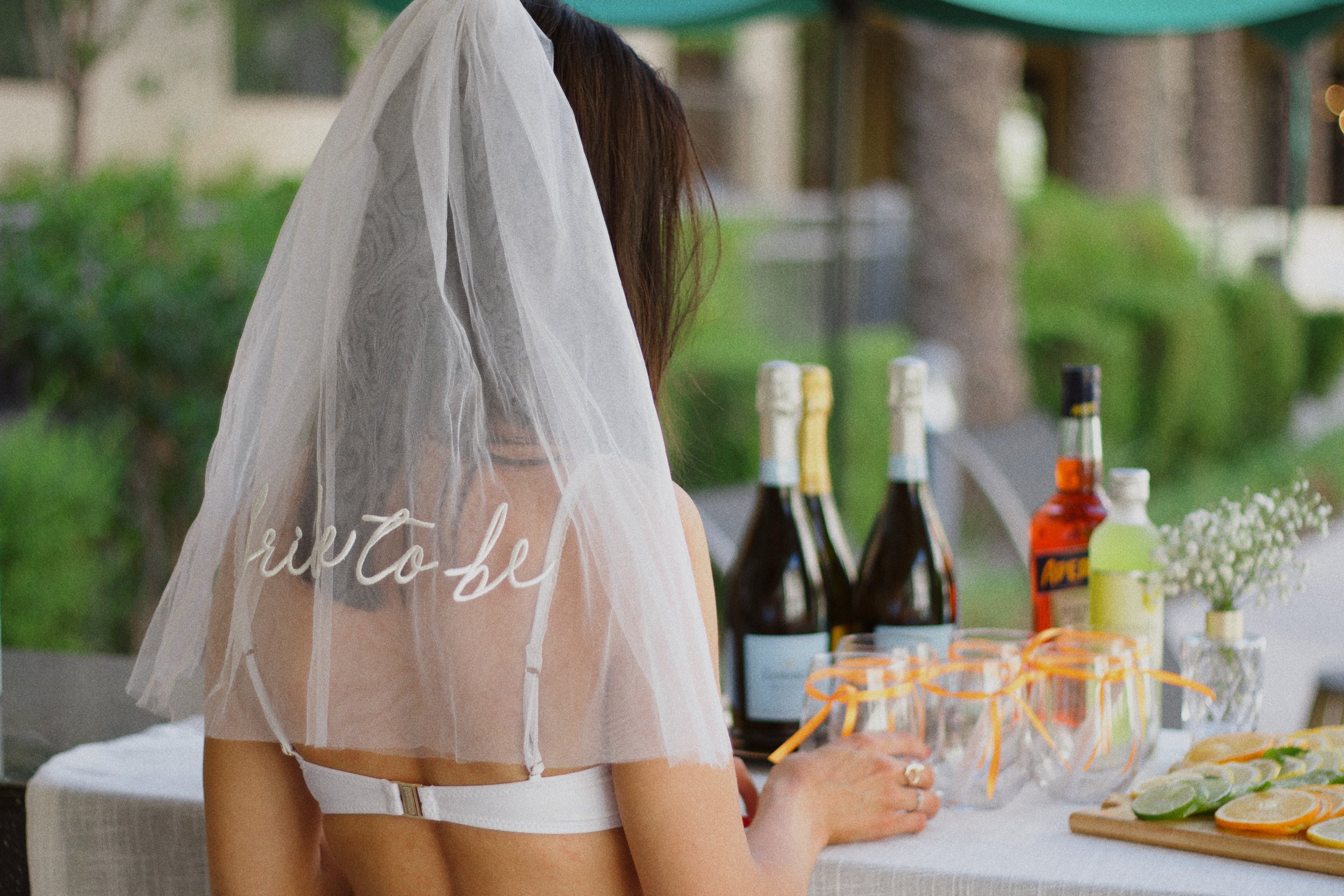 A woman wearing a wedding veil and white swim suit at a table outdoors, with bottles of alcohol, glassware, and sliced citrus on a wooden cutting board. It is a very appealing set up and great for outdoor get togethers.