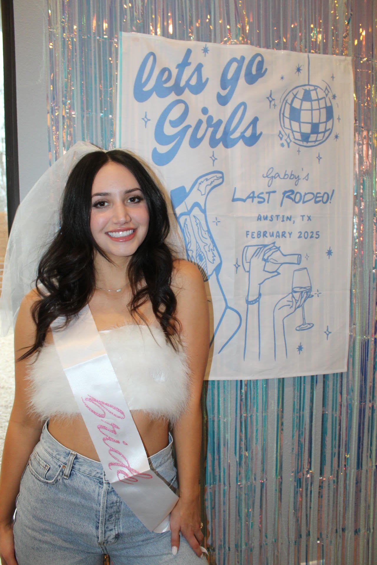 A young woman at a celebration wearing a white sash and fluffy top, standing in front of a sign that reads 'Let's go girls' with blue text and illustrations of a hand holding a microphone and a wine glass, in front of a shiny, iridescent backdrop.