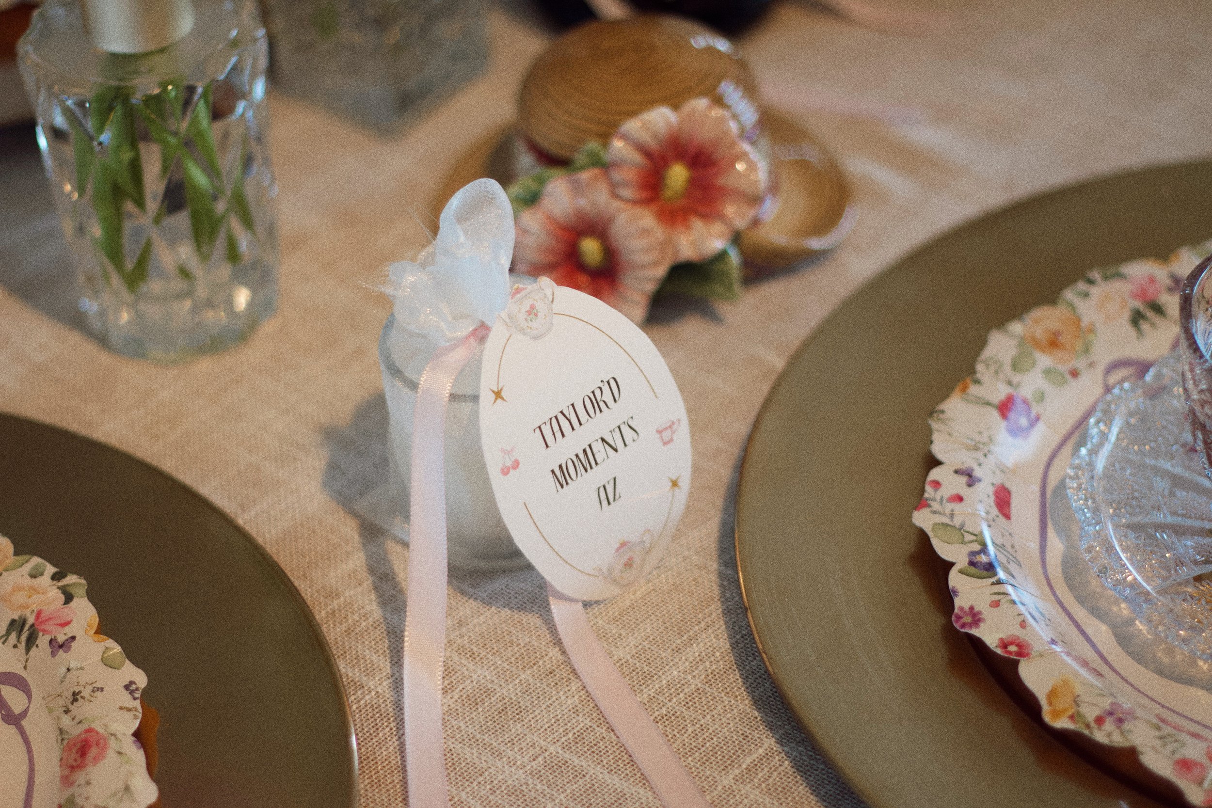 A decorated table setting for a celebration with floral-themed napkins, gold plates, a small bottle with a butterfly and ribbon, and a circular tag with flowers and the text 'Taylor'd Moments AZ', placed on a beige tablecloth.