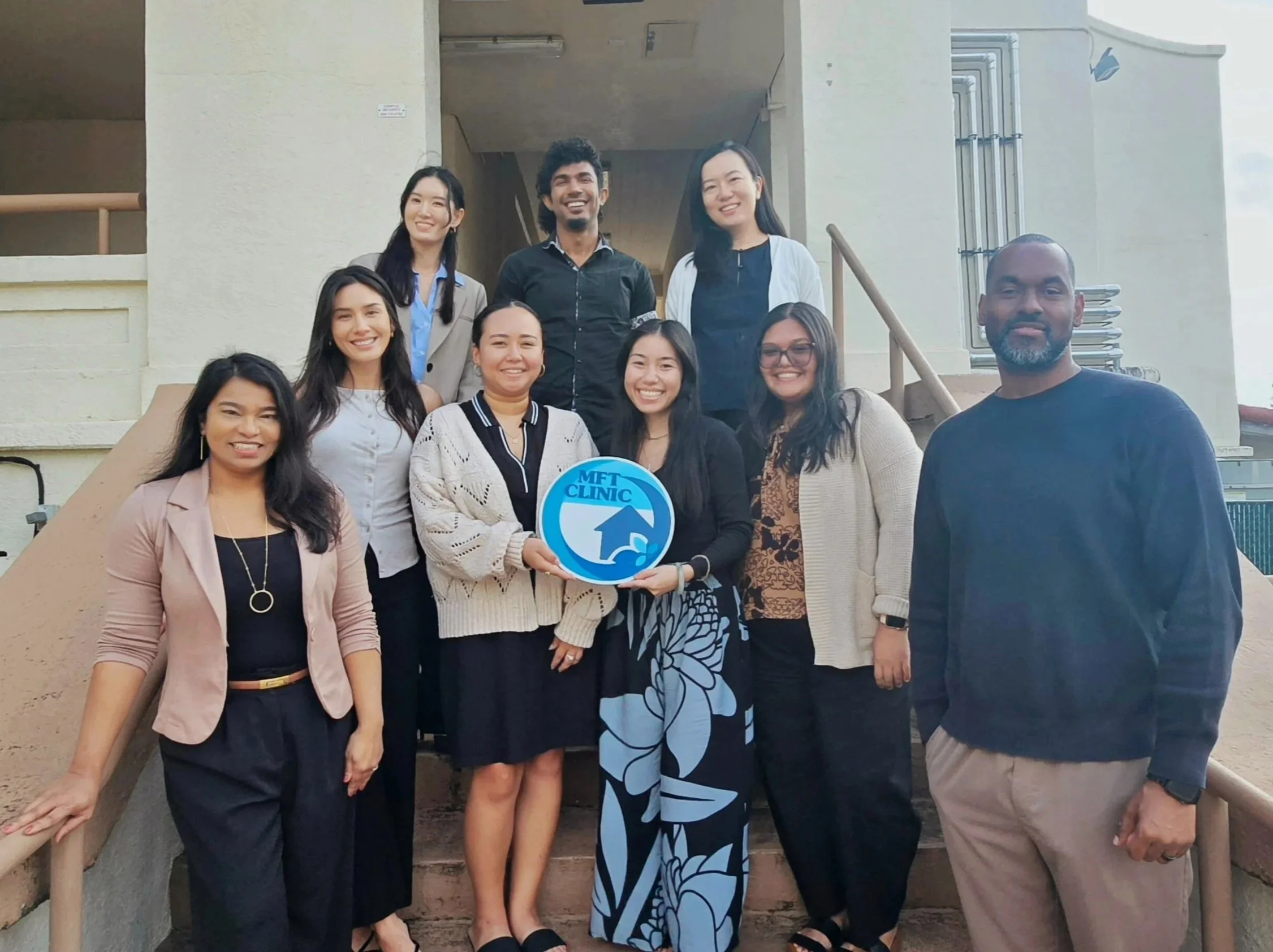 Group of ten Group of eleven diverse Chaminade University MS-MFT Graduate student clinicians and clinic faculty/staff  standing on stairs outside, holding a blue sign that says 'MFT CLINIC', smiling for the camera.