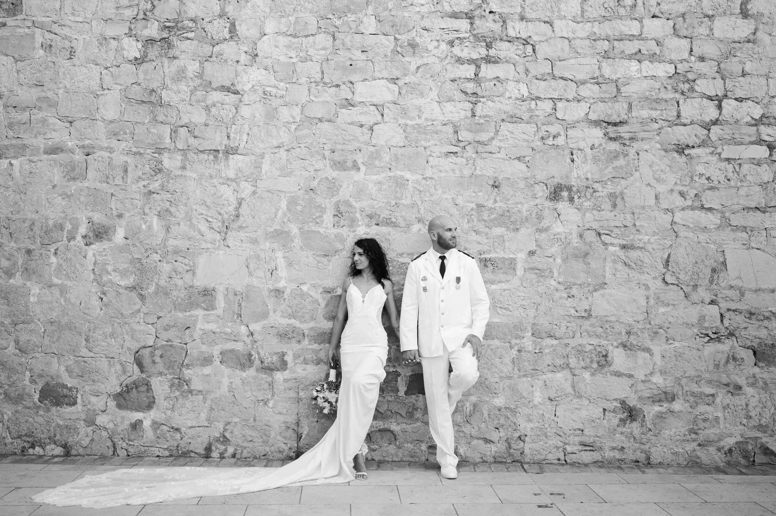 Couple de mariés habillés en blanc, adossés contre un mur de pierres Provençales