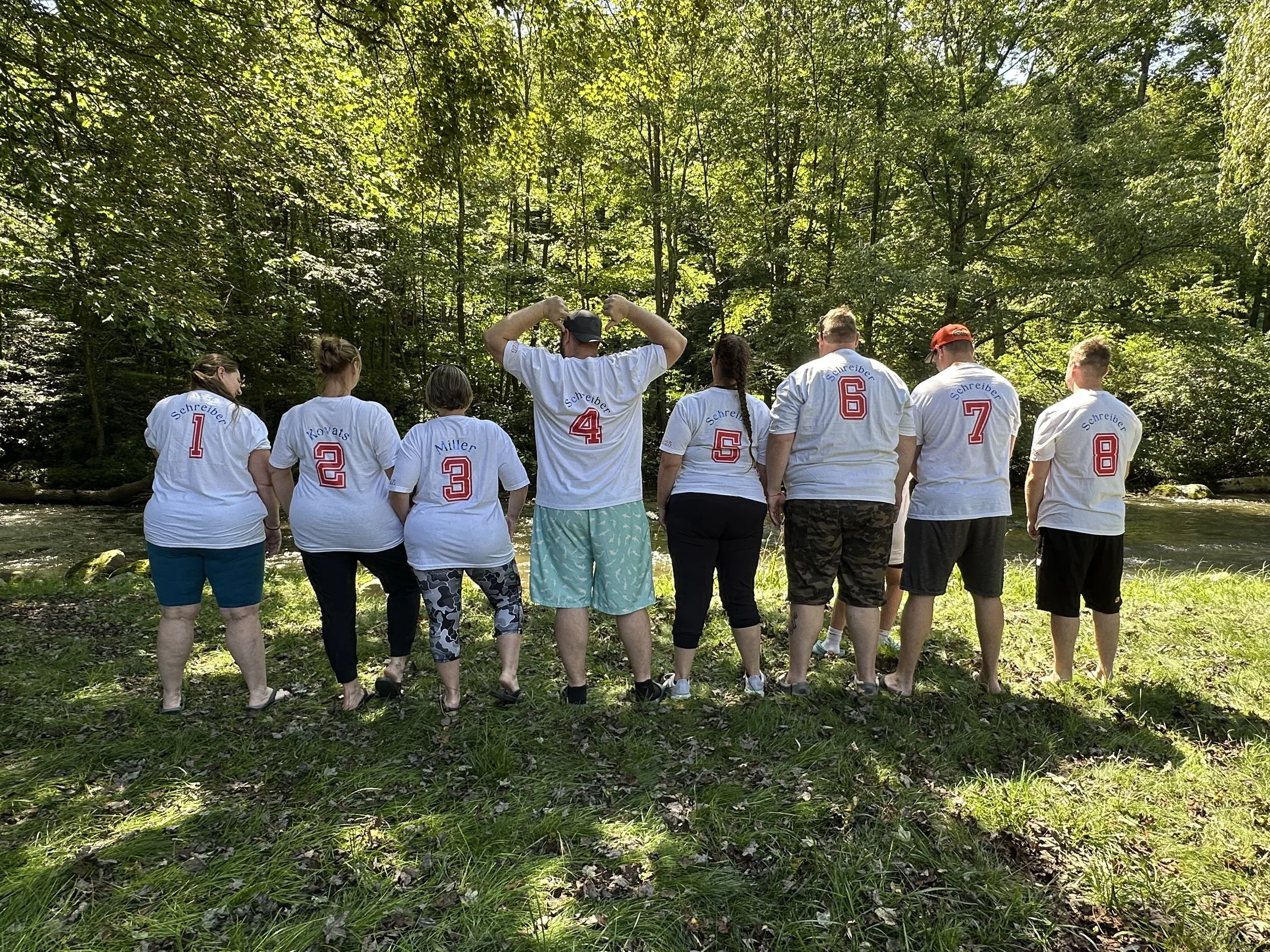 A group of nine people standing outdoors near a stream, wearing matching white T-shirts with red numbers on the back, in a wooded area.