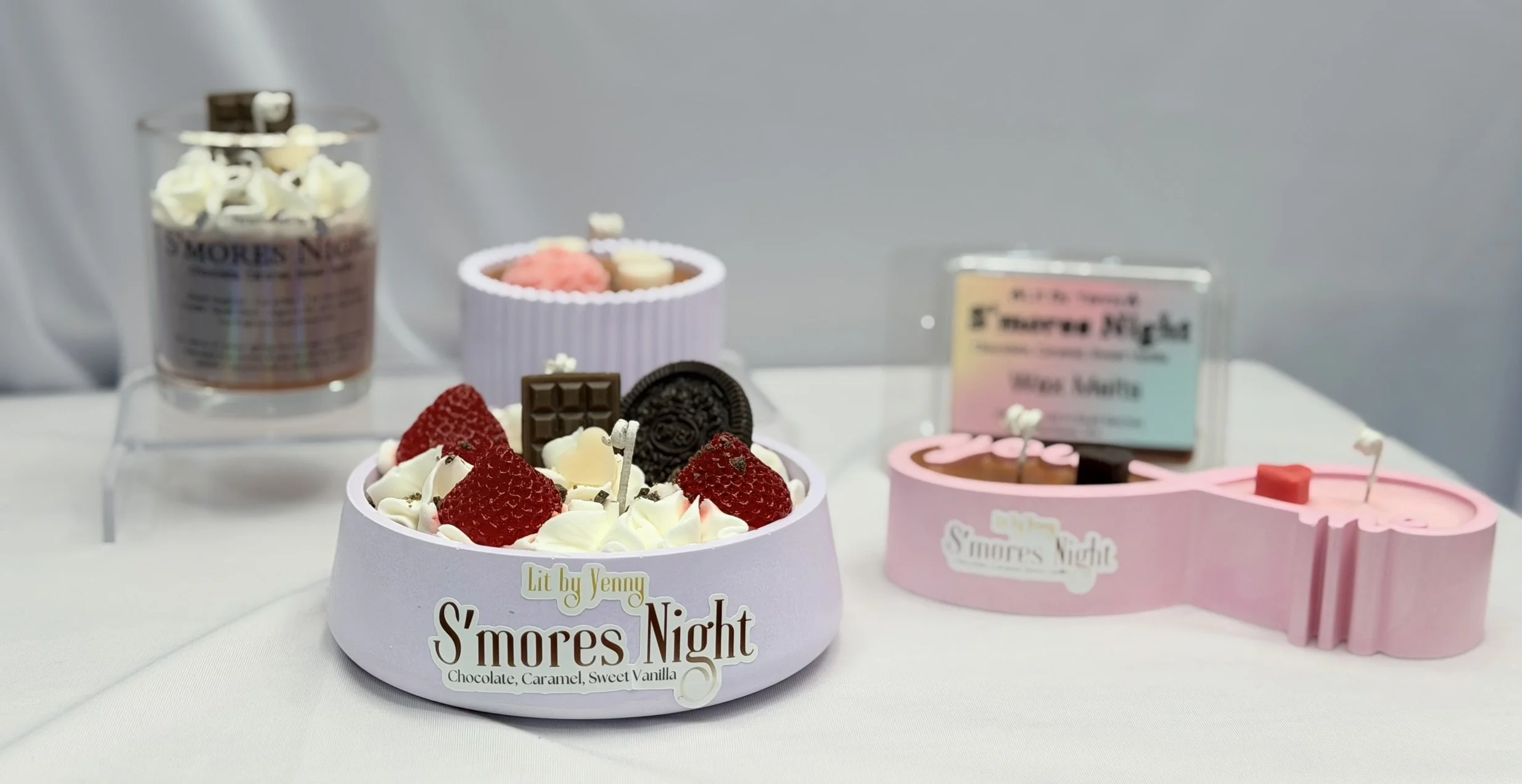 Table display with assorted treats including a pink and purple mini guitar-shaped cake with whipped cream and strawberries, a group of chocolates and cookies, several small jars of candy, and a sign with the label "S'mores Night".
