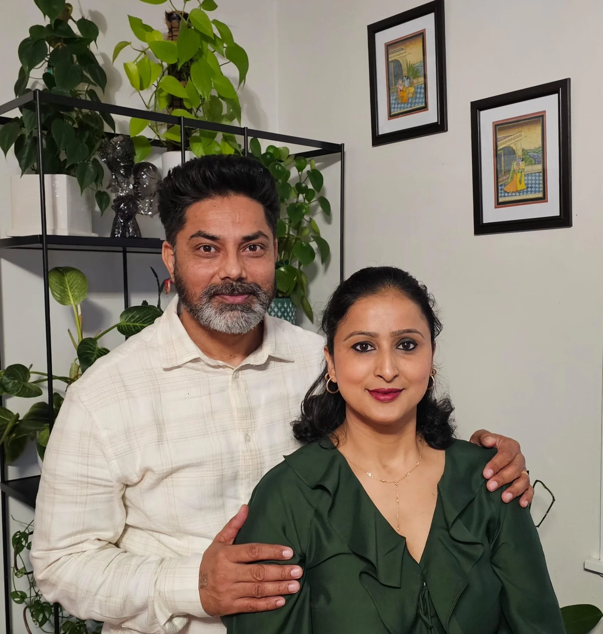A man and woman standing close together indoors, with the man’s arm around the woman’s shoulder. The man has grey hair, a beard, and is wearing a light beige shirt. The woman has dark hair styled back, and is wearing a dark green blouse and gold jewelry. Behind them is a black metal shelf with green houseplants, and on the wall, two framed pictures with colorful scenes.