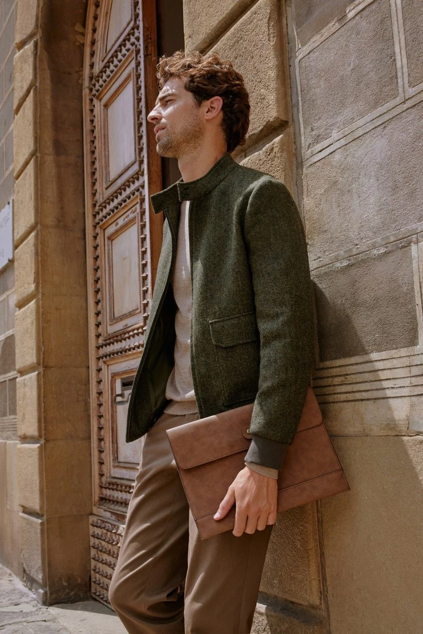 Man with curly hair wearing a green jacket and beige pants, holding a tan leather folder, standing against a stone wall in an urban setting.