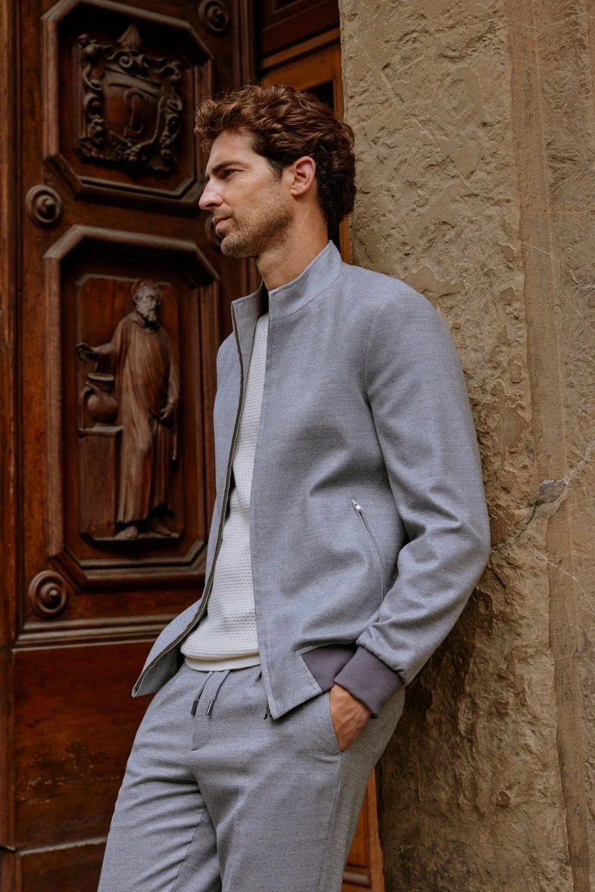 A man with brown curly hair and a beard leaning against a beige stone wall and wooden door with carved details and religious figures.