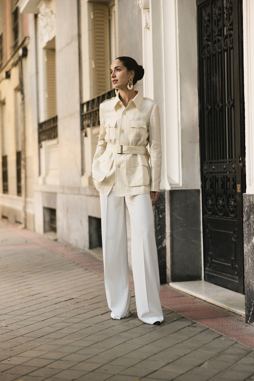 Woman in a beige jacket and wide-leg white pants standing on a city sidewalk in front of an ornate black and white building.