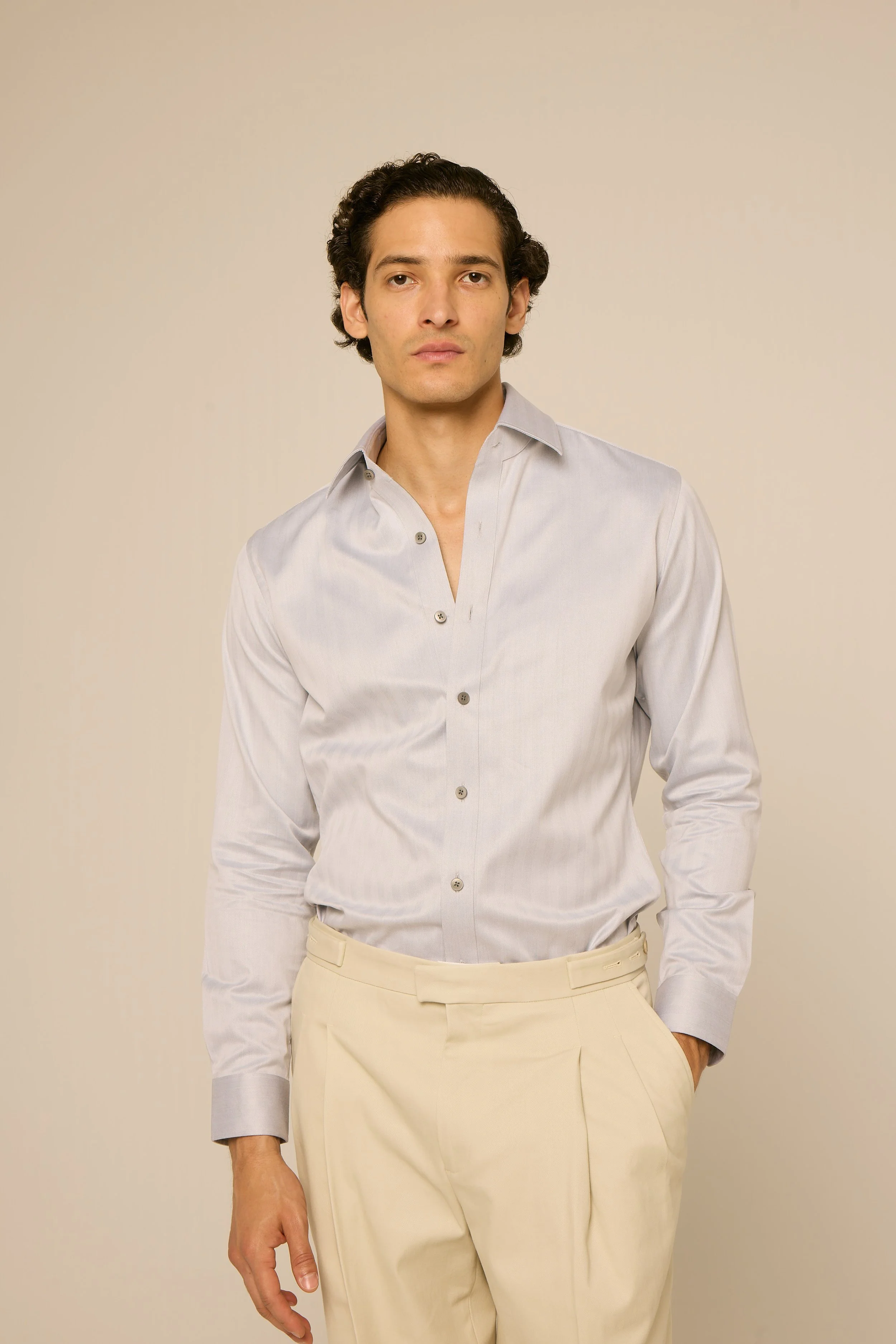 A young man with dark, curly hair wearing a light-colored button-up shirt and cream-colored pants, standing against a plain, neutral background.