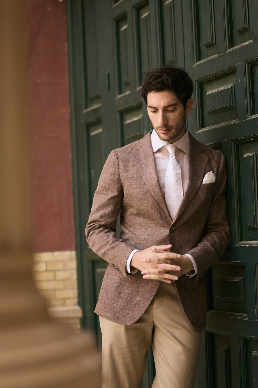A young man in a brown blazer, white shirt, and tie standing with closed eyes and hands clasped in front of green paneling and a brick wall.