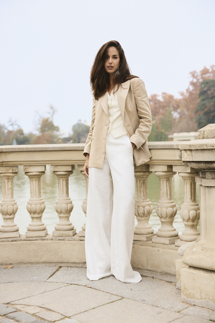 A woman in a beige blazer and wide-legged white pants standing on a stone balcony with ornate balustrades near a body of water, with trees and an overcast sky in the background.