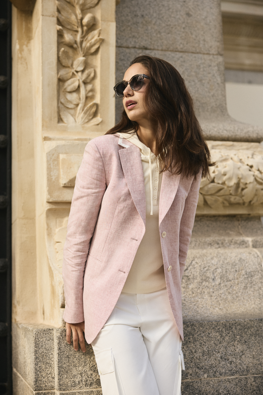 A woman with brown hair wearing sunglasses, a beige blouse, a pink blazer, and white pants, leaning against a decorative stone structure outside.