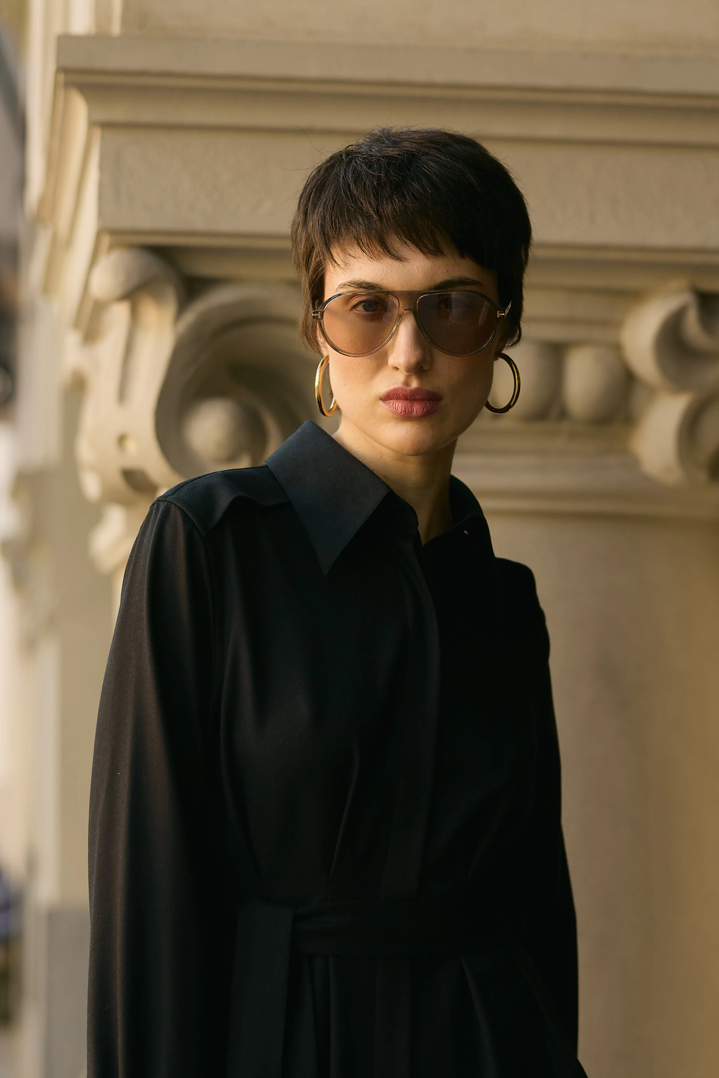 A woman wearing large sunglasses and gold hoop earrings, dressed in a black outfit with a belt, standing outdoors in front of decorative architecture.