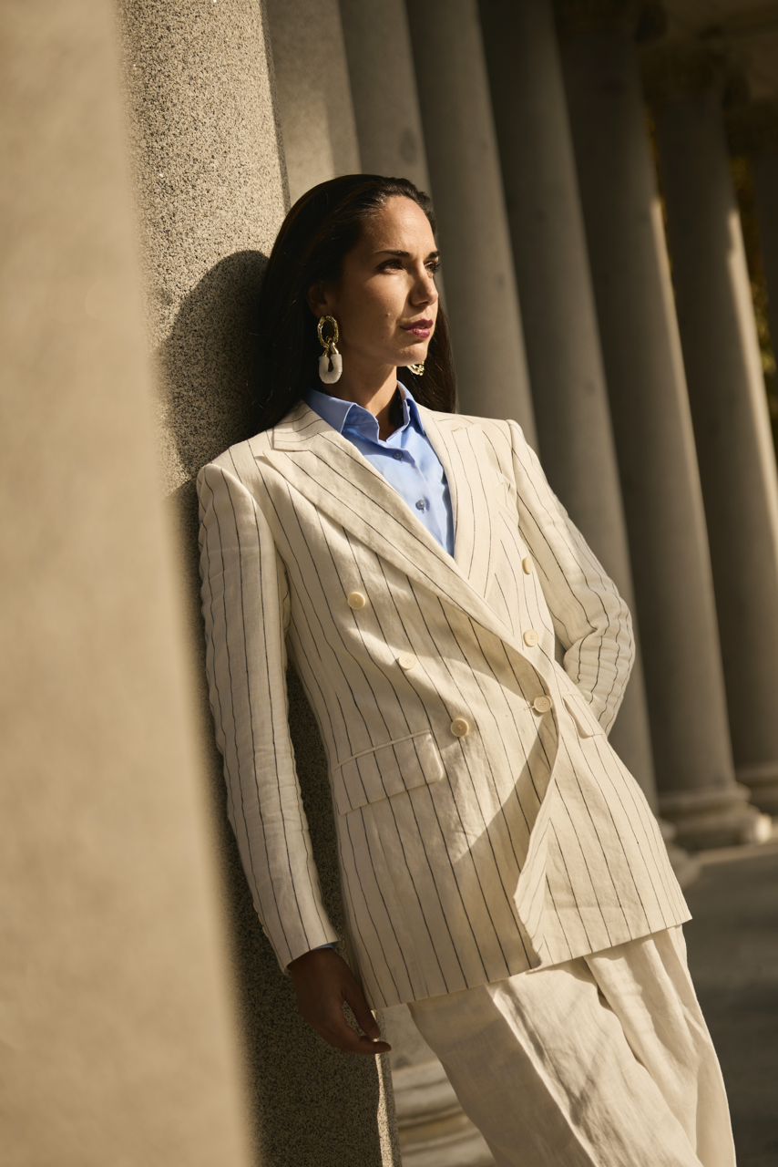 A woman dressed in a cream pinstripe blazer and light blue shirt leaning against a textured stone column during sunset.