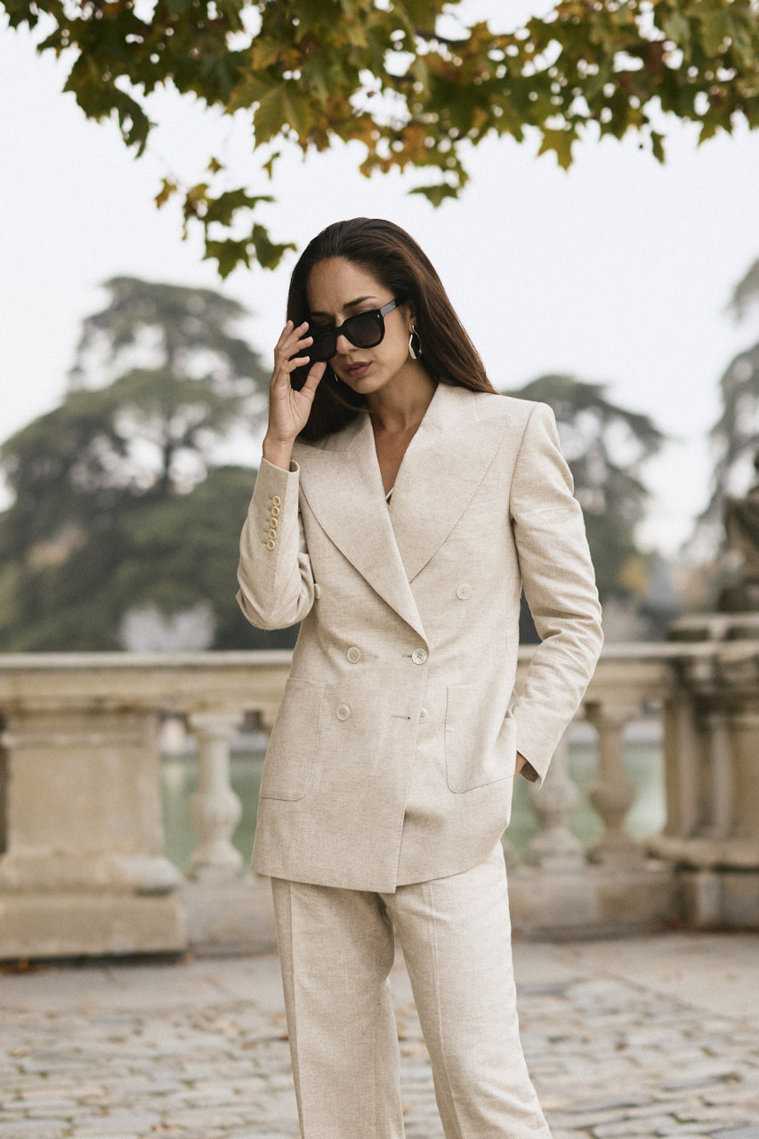 A woman in a beige double-breasted blazer and matching pants, wearing dark sunglasses, standing outdoors on a stone-paved area with a stone balustrade and trees in the background.