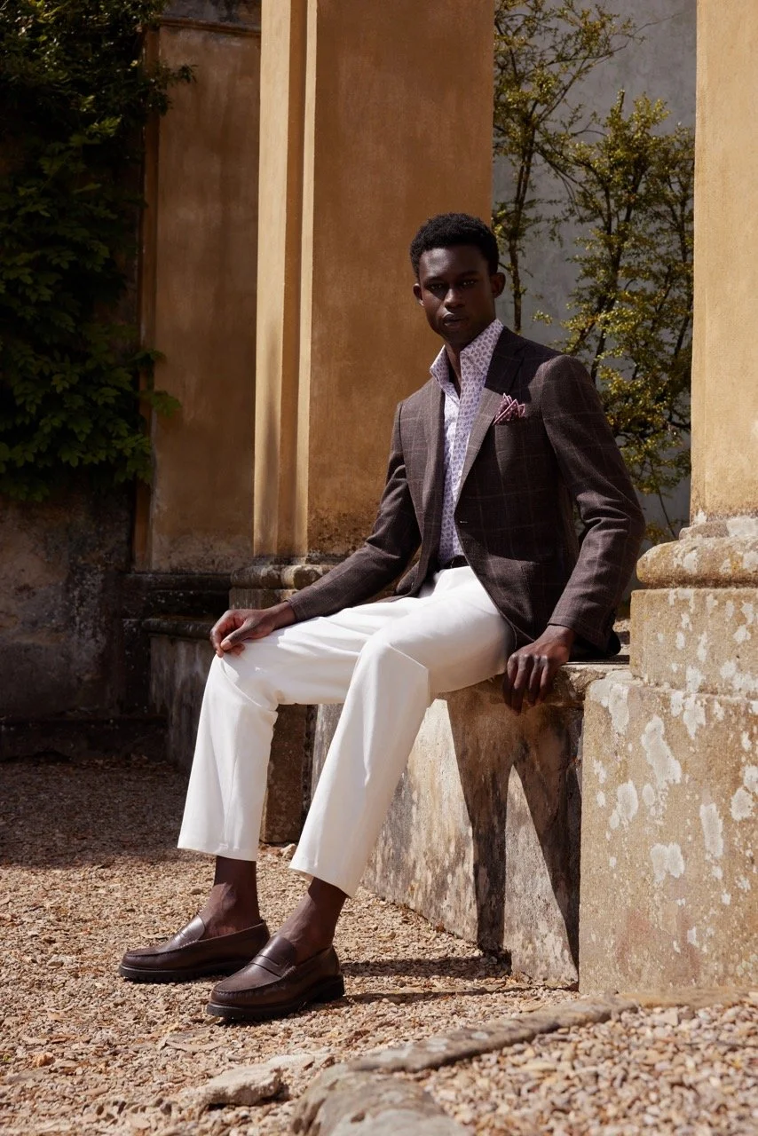 A young man is sitting on a stone bench outdoors, dressed in a brown checked blazer, white trousers, a patterned shirt, and brown loafers, with trees and a historic wall in the background.