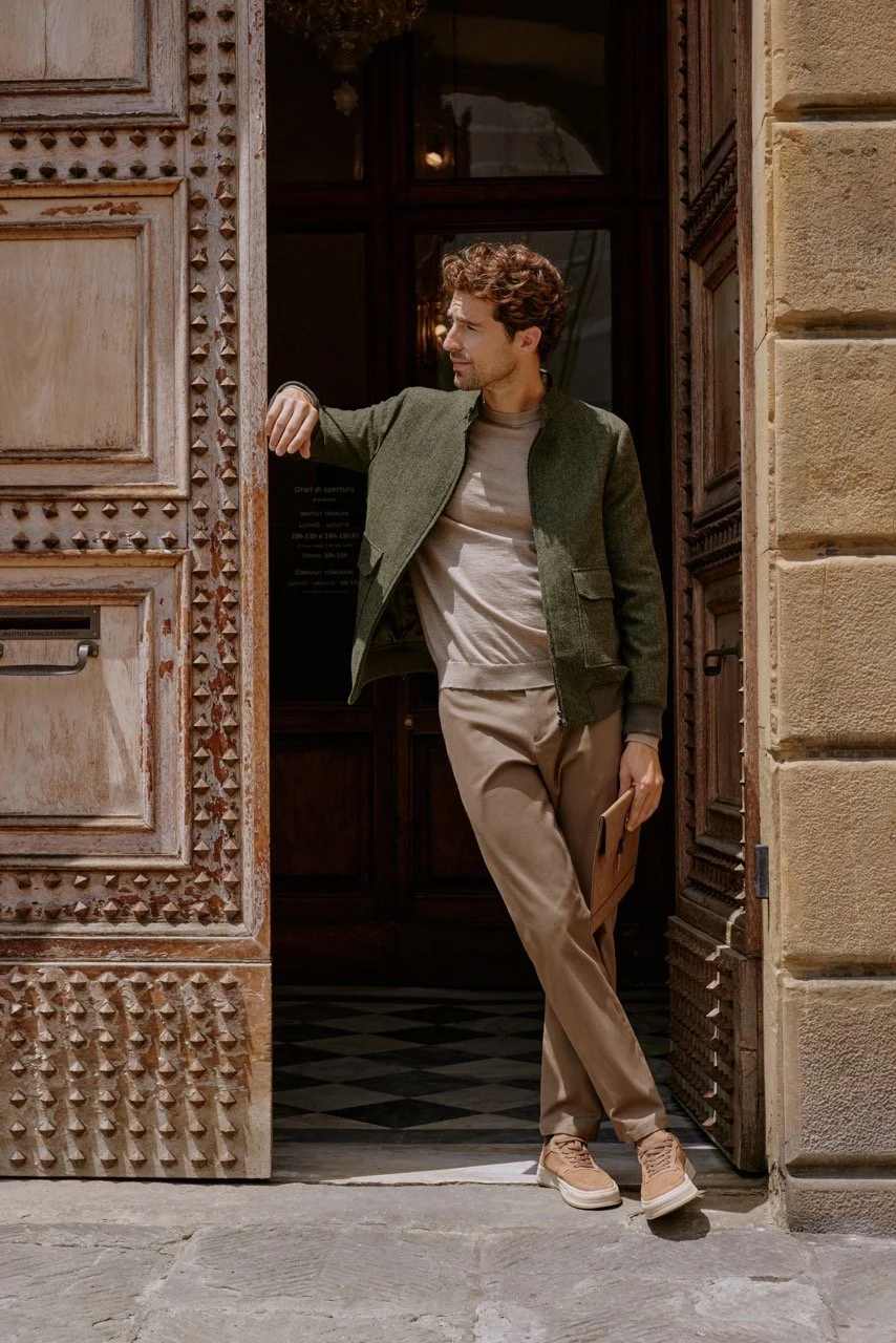 Young man standing at an open doorway, wearing beige pants, light brown sneakers, a beige shirt, and a green jacket, with a brown folder in his hand, looking to the side.