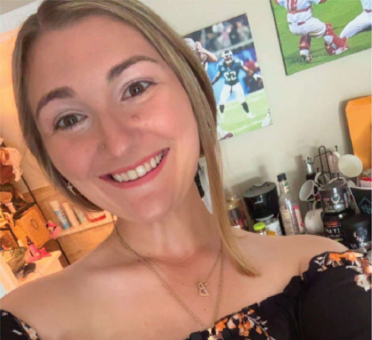 A smiling woman with shoulder-length blonde hair, wearing a black off-the-shoulder top with floral pattern, in a room decorated with sports posters, kitchen items, and shelves.