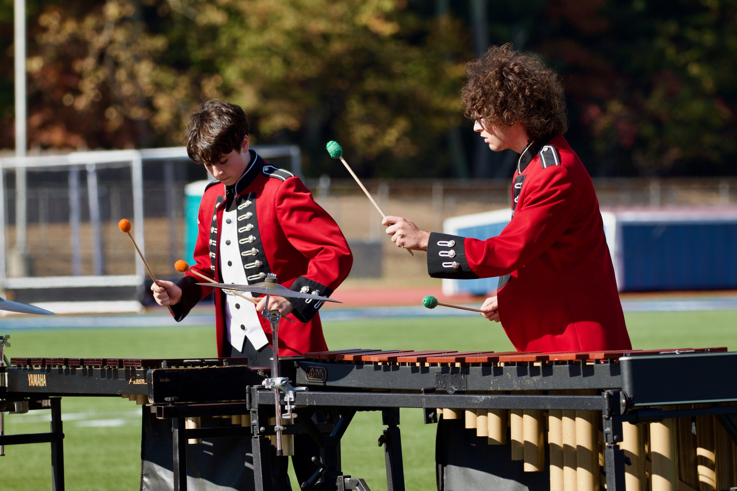 Two young musicians dressed in red and black marching band uniforms playing percussion instruments outdoors.
