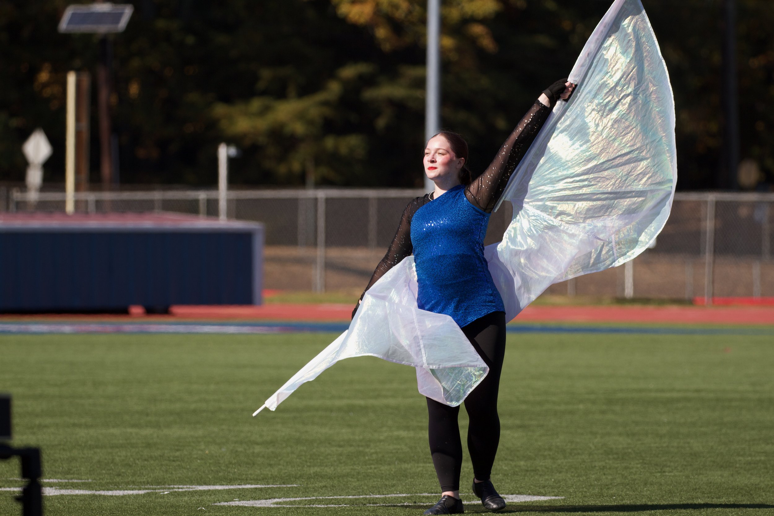 A young woman in a blue sequined top and black leggings performing a flag dance on a football field with a large iridescent flag.