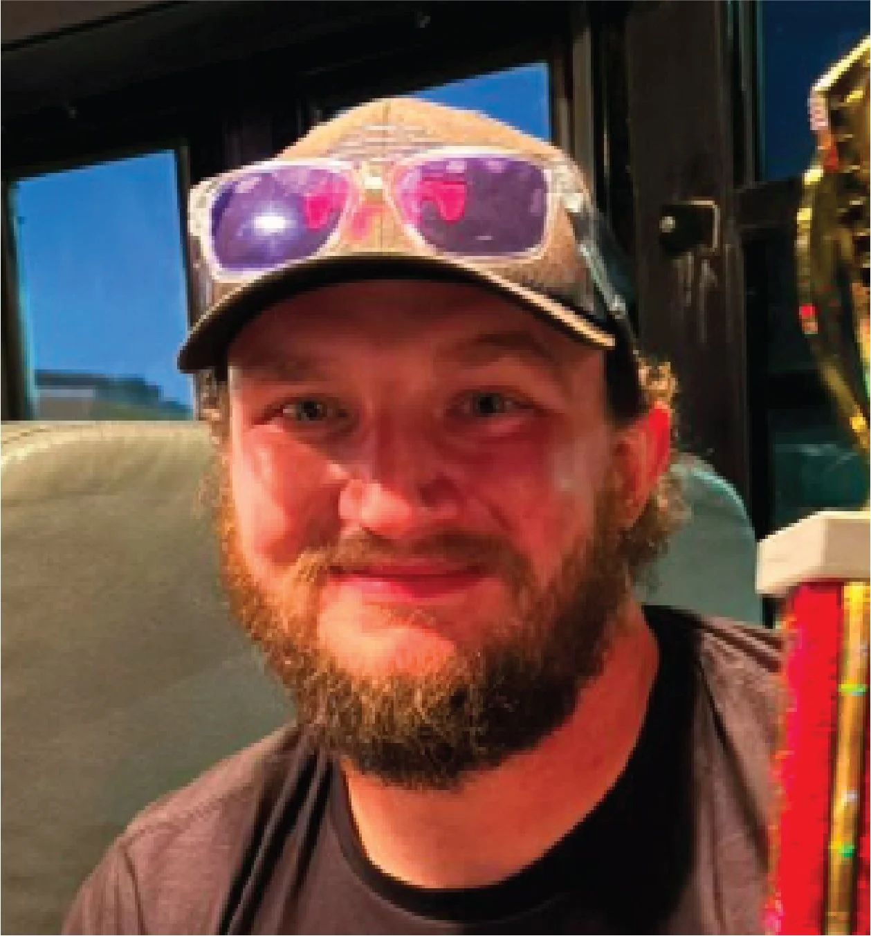 A smiling man with a beard wearing a tan cap with purple sunglasses on top. He has on a black T-shirt and is sitting inside a vehicle, likely a bus.