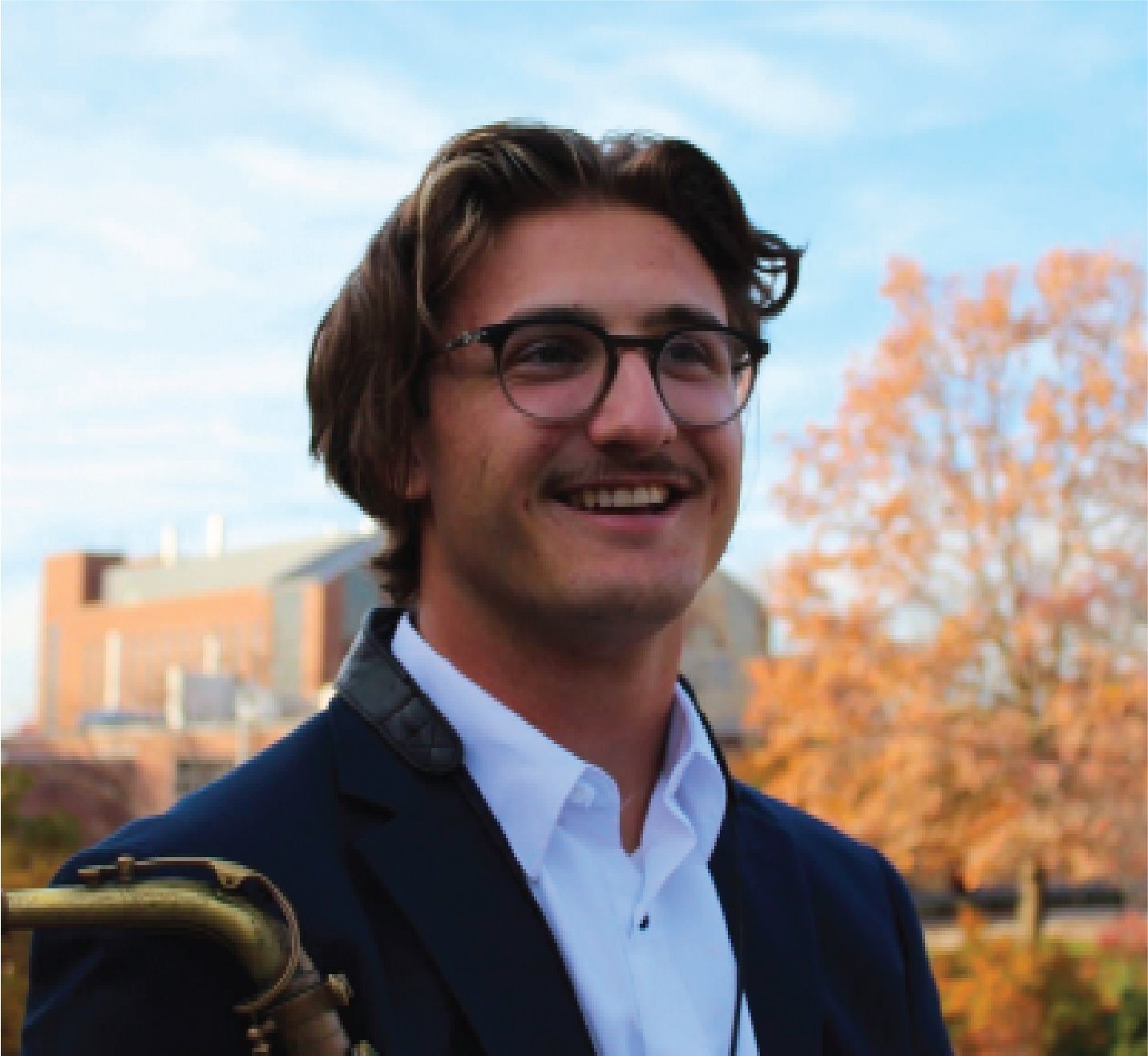 A young man with brown hair, glasses, and a mustache in an outdoor setting during autumn, smiling and wearing a dark blazer and white shirt.