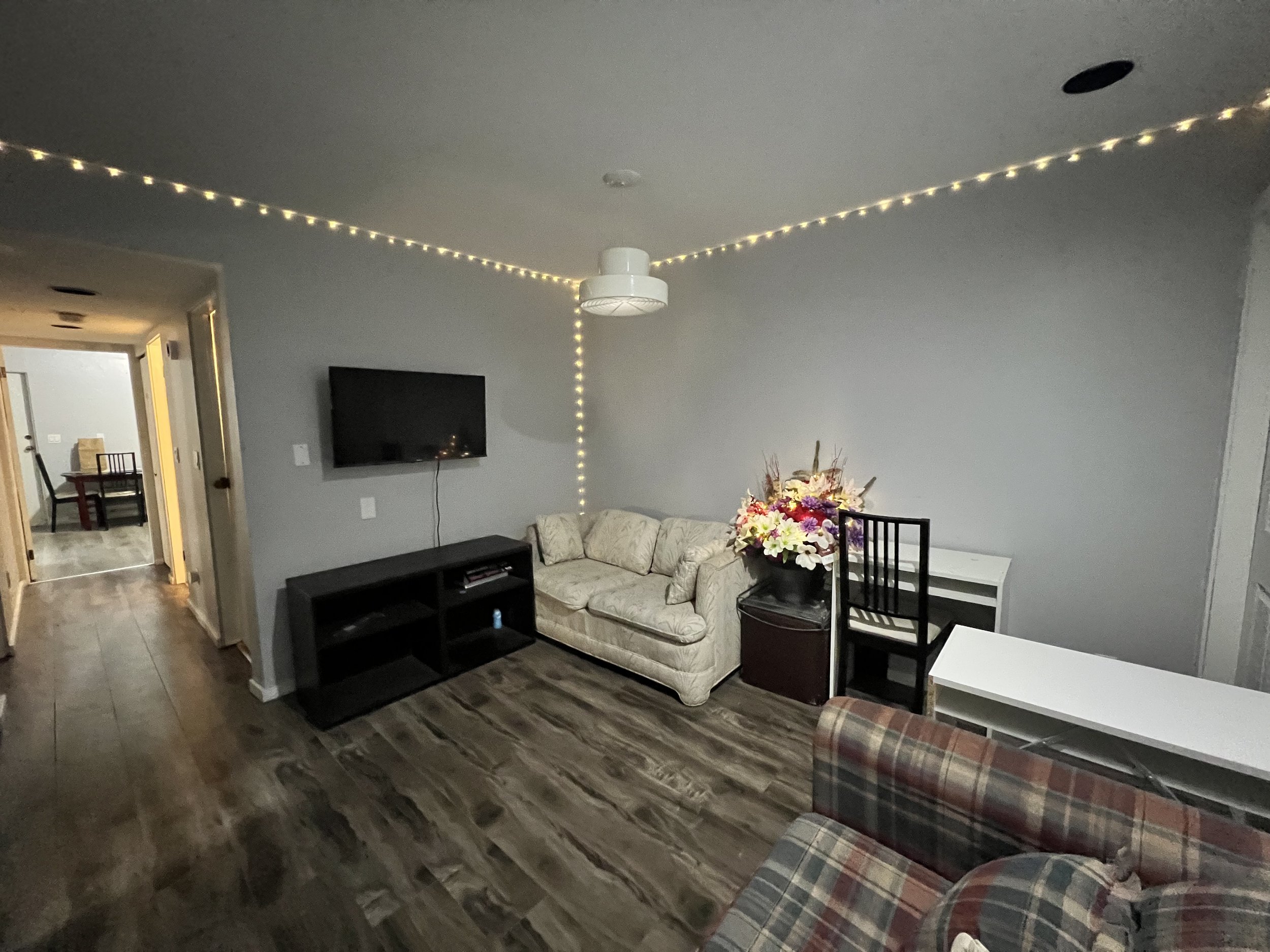 Living room with a beige sofa, a black entertainment center, a wall-mounted TV, and a white desk with a black chair. There's a vase of flowers on a side table, and string lights hanging from the ceiling around the room. The floor is wood and there's 