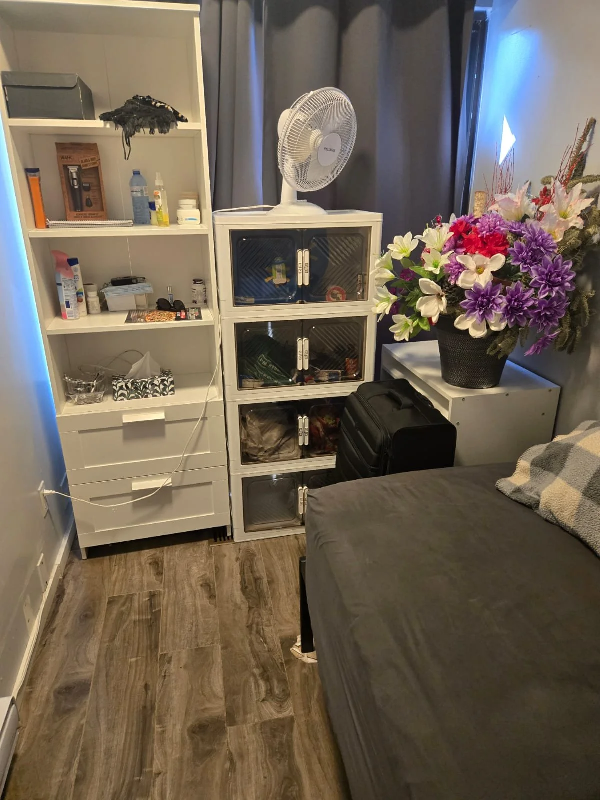 A bedroom corner with a white shelving unit, a fan, two small white cabinets, a black suitcase, a gray bed with a checkered pillow, and a large bouquet of pink, purple, and white flowers.