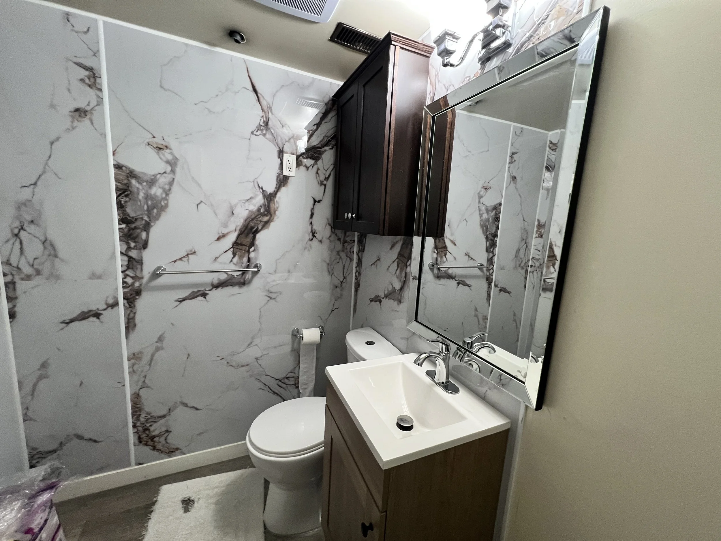 Bathroom with marble-patterned wall, a white toilet, a small white sink with a cabinet, a large rectangular mirror, their dark wall cabinet, and part of a towel rack, with a beige wall on the right side.