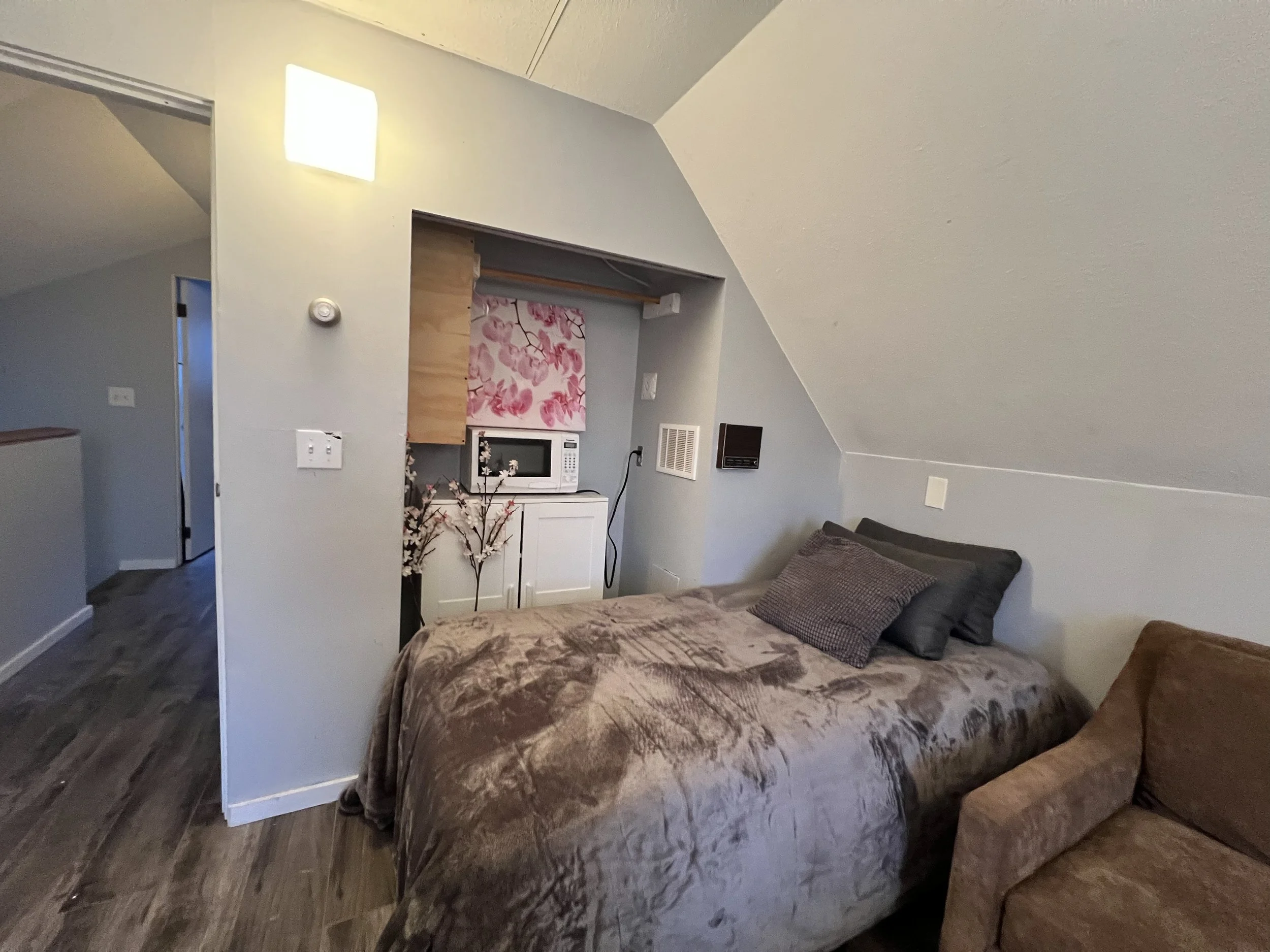 A cozy bedroom with a slanted ceiling, a bed with brown velvet bedding, pillows, and a small sitting area with a brown chair. A kitchenette area with a microwave, cabinets, and decorative pink flower artwork is visible.