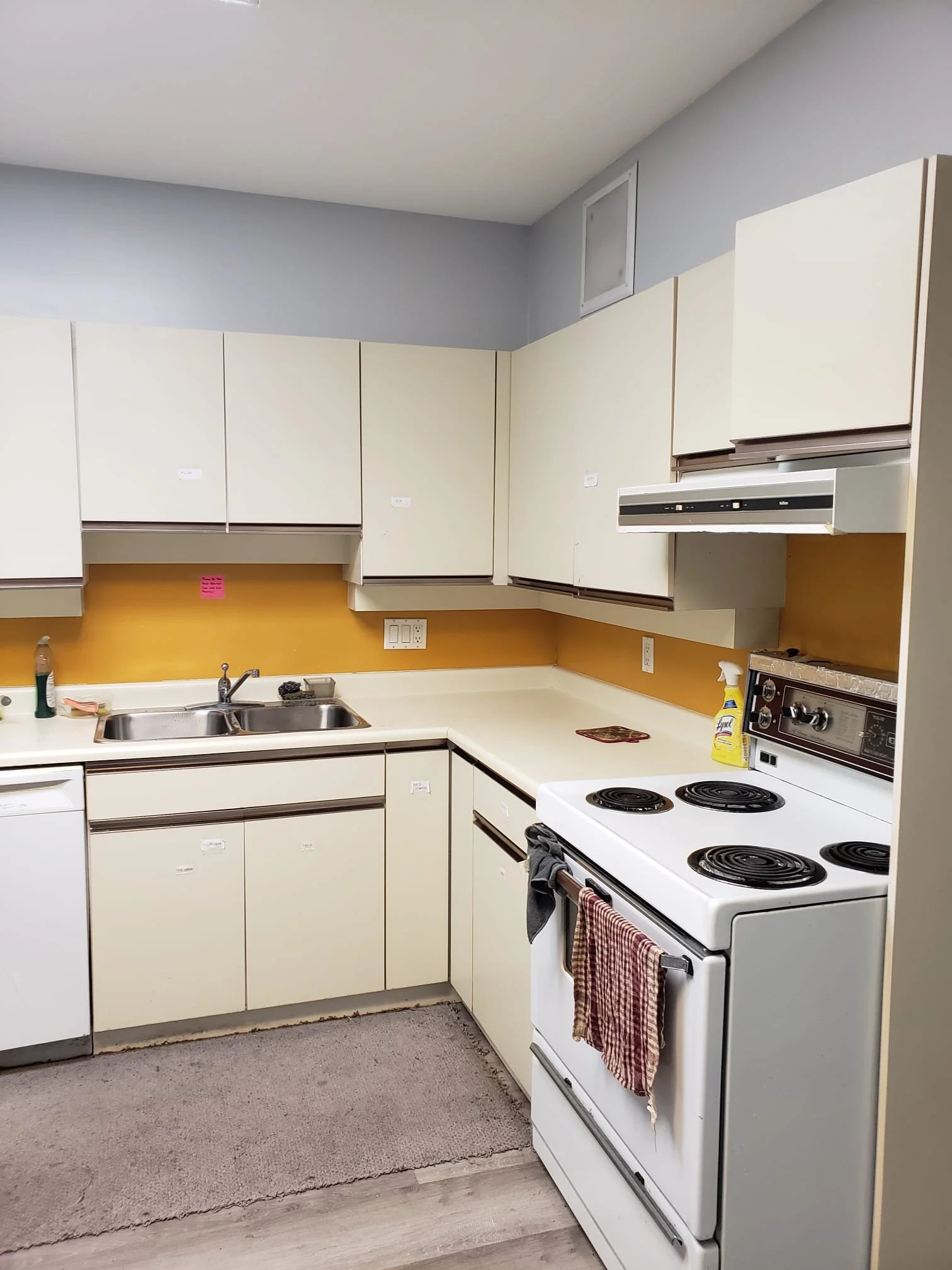 An old kitchen with white cabinets, a yellow backsplash, a double sink, and a white electric stove with towels hanging on the oven handle.