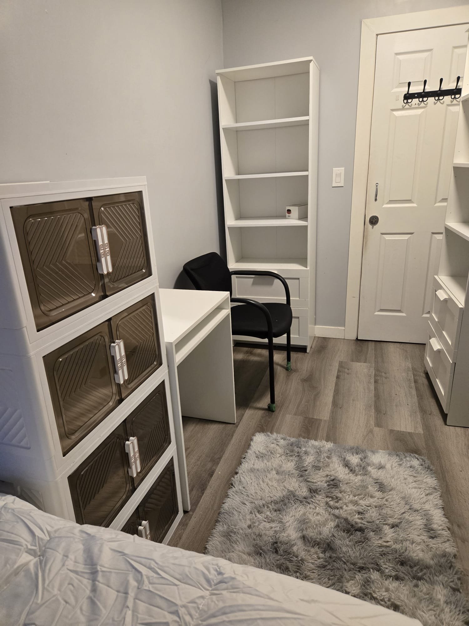 A small bedroom or office space with white furniture, including a tall white bookshelf with three shelves and a lower cabinet, a white desk, and a white storage cabinet with brown tinted glass doors. There is a black chair with armrests, a small fluf