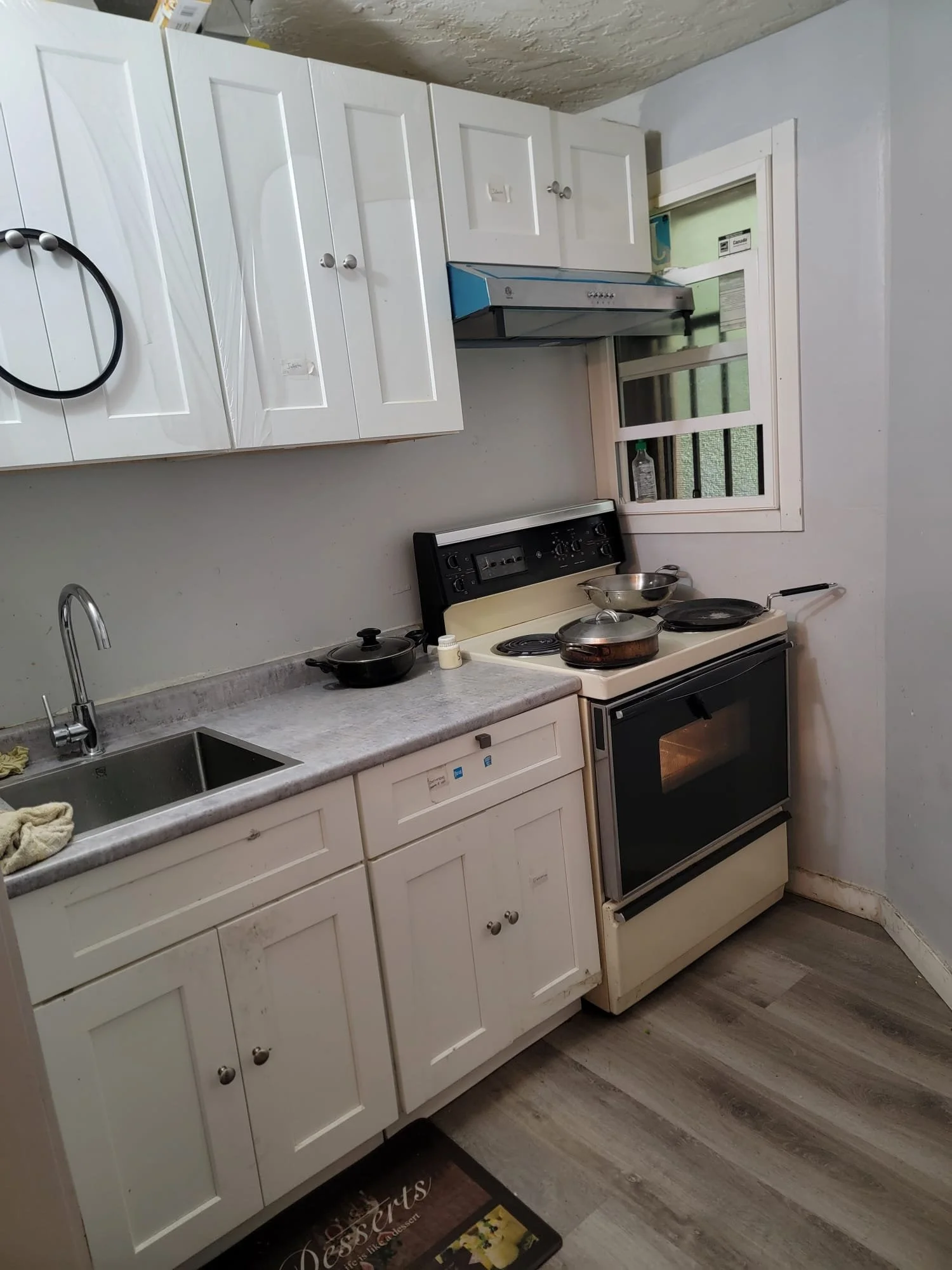 A small kitchen with white cabinets, a gray countertop, a stainless steel sink, a stove with four burners, a window above the stove, and a range hood. There are pots and pans on the stove and countertop.