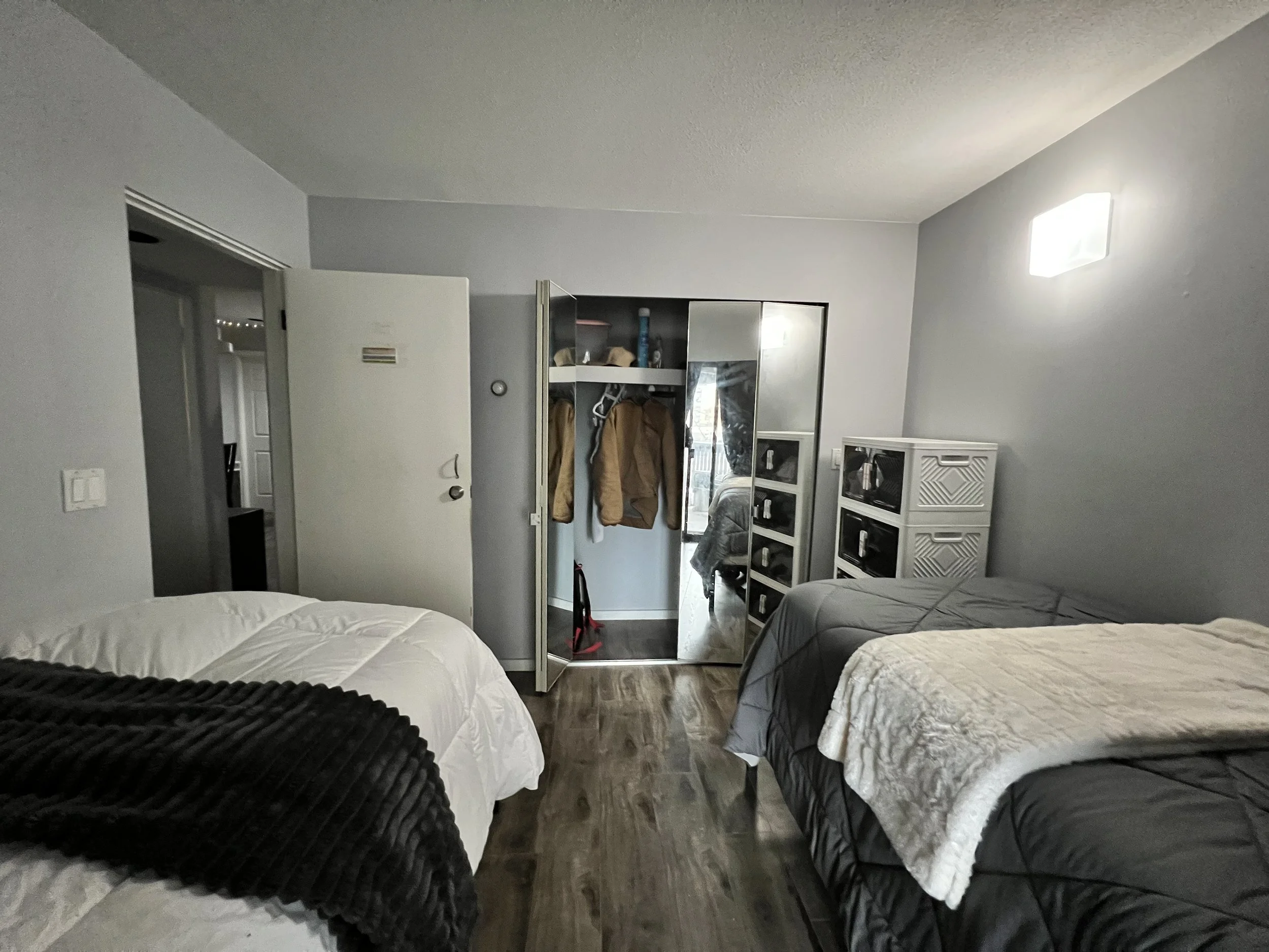 A bedroom with three beds, one with white bedding and black throw, another with gray bedding, and the third with beige bedding. A closet with a mirror door is open, showing jackets and other items inside. There are storage cubes on the right side of 