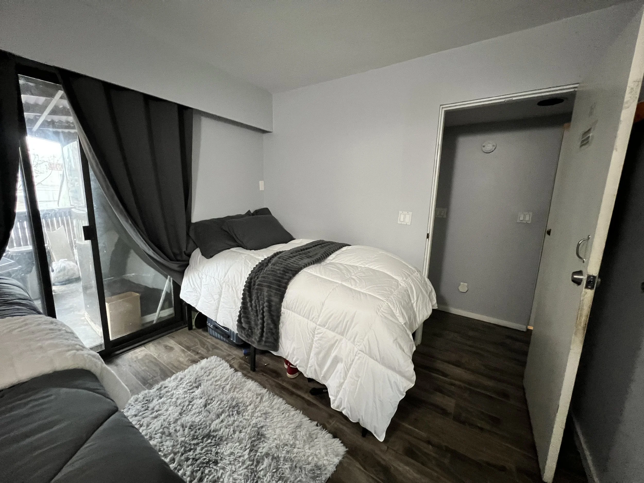 Small bedroom with a bed covered in white bedding and black pillows, a dark gray throw blanket, black curtains over a sliding glass door, and a plush gray rug on a dark wood floor. The room has gray walls and an open door leading to a hallway.
