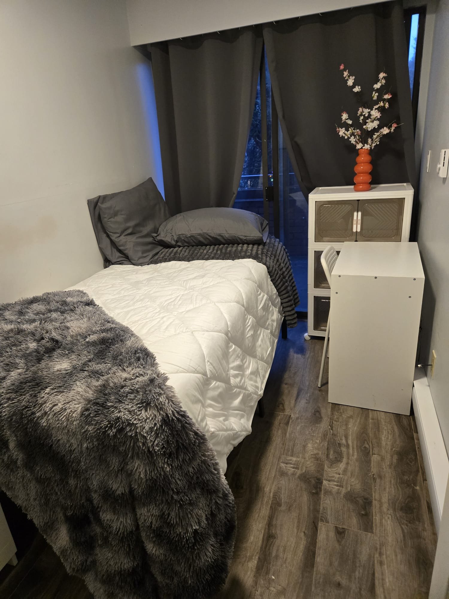 Small bedroom with a bed on the left side, covered with gray pillows, a gray blanket, and a faux fur throw. A nightstand or small cabinet is in the corner with a tall orange vase holding pink and white flowers. The room has dark curtains and a sliding window or door behind them, with a view of trees outside.
