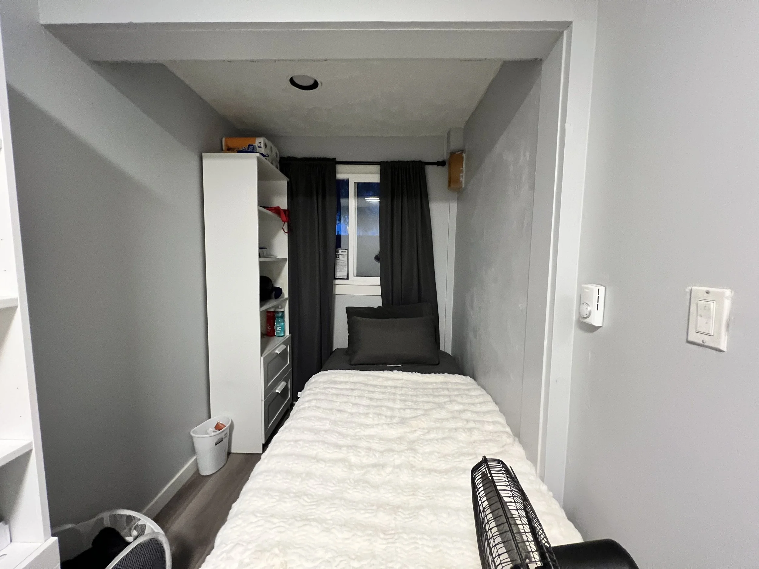 A small bedroom with a single bed covered with a white fluffy blanket, two dark pillows, a small window with dark curtains, a white shelf with items, a small trash bin, and a portable fan.